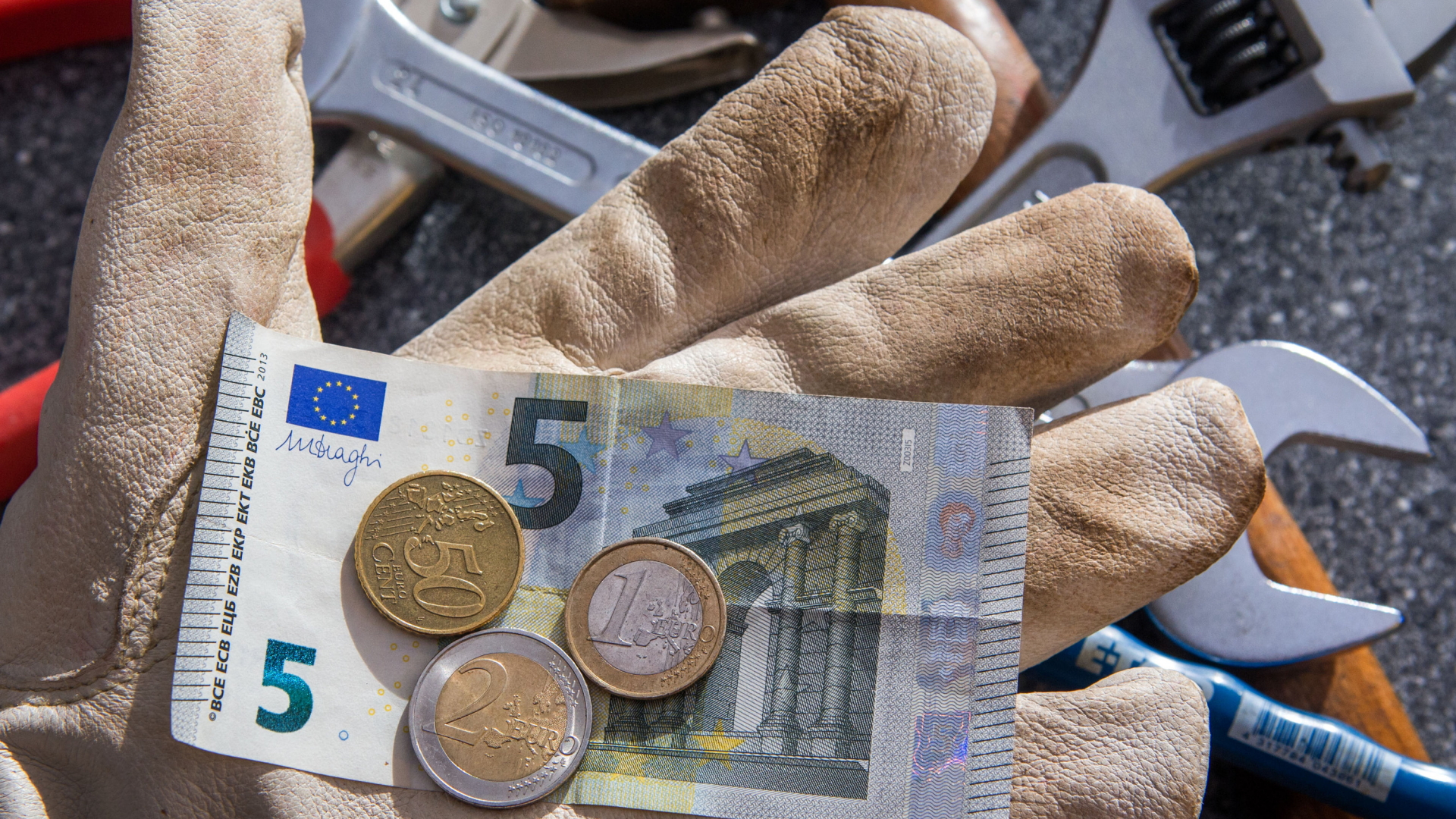 Ein Handwerker mit Arbeitshandschuh hält einen Fünf-Euro-Schein und ein paar Münzen in der Hand.