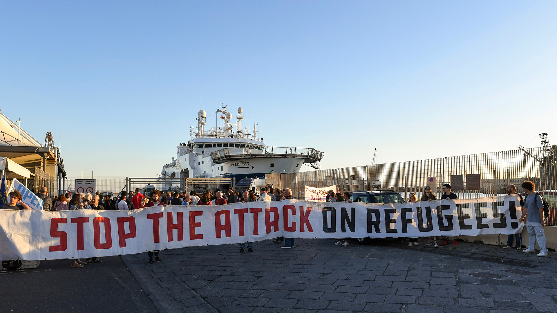 Demonstranten halten ein Transparent mit der Aufschrift "Stop the Attack on Refugees" (Stoppt den Angriff auf Flüchtlinge) vor dem Rettungsschiff  "Geo Barents" im Hafen von Catania.  | dpa
