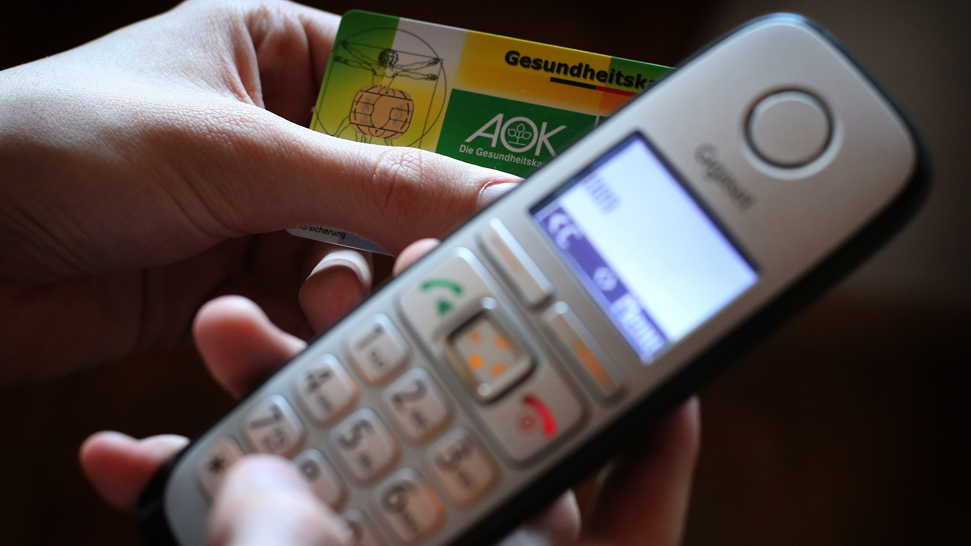 Ein Mann hält eine Gesundheitskarte der Allgemeinen Ortskrankenkasse (AOK) in der Hand, während er am Telefon eine Nummer wählt.