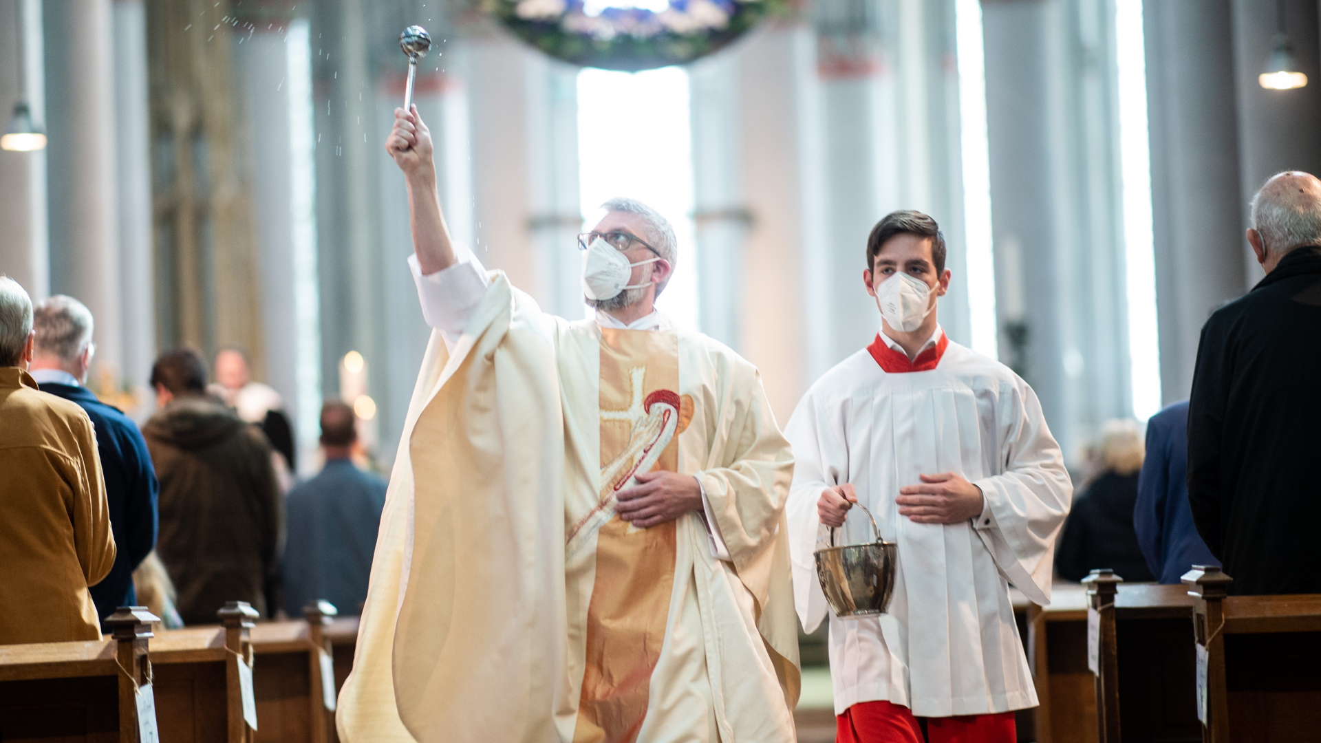 Ein Pfarrer mit Maske teilt Wasser auf die Gläubigen zur Erinnerung an die Taufe aus. | dpa