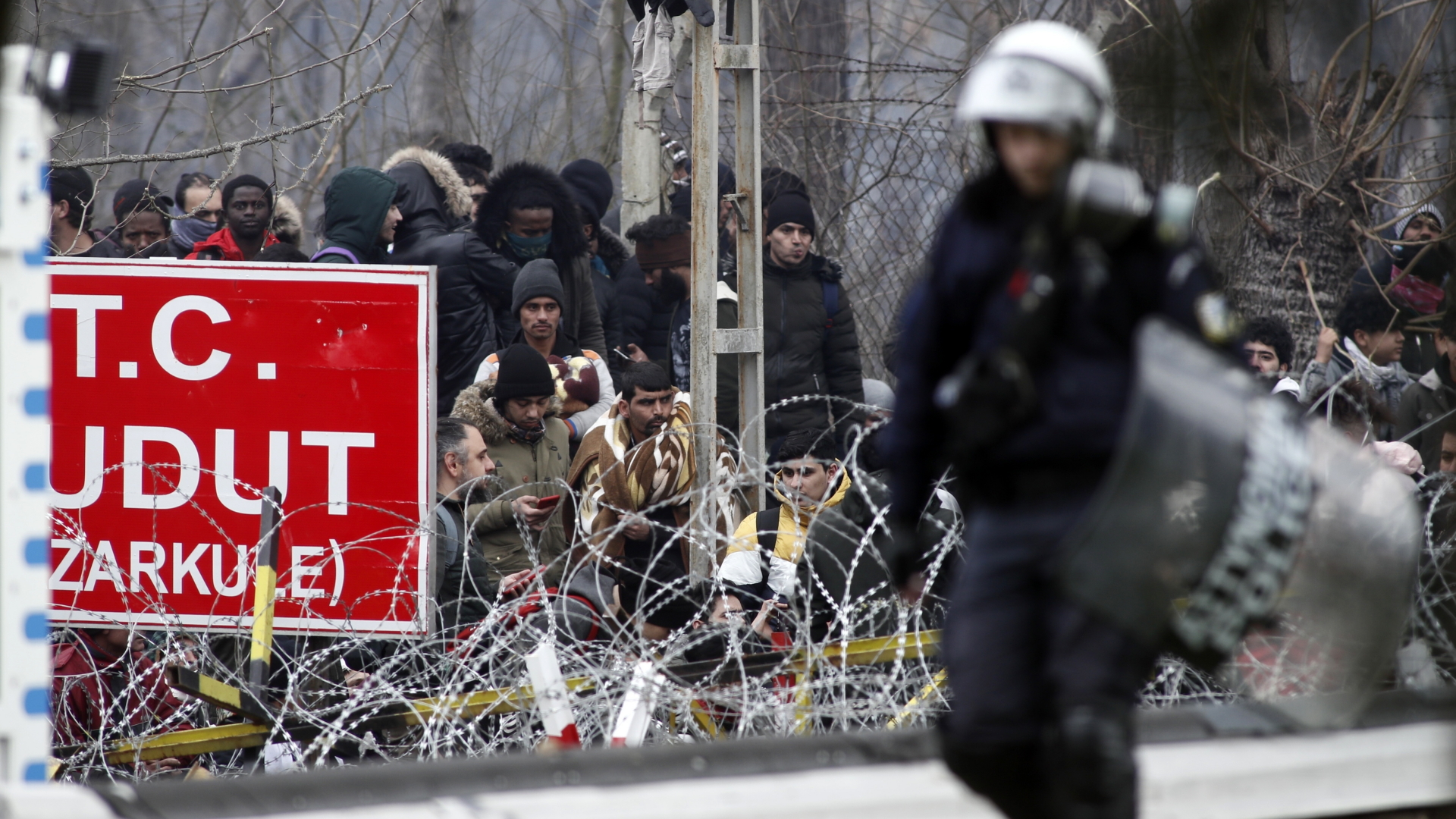 Ein griechischer Bereitschaftspolizist patrouilliert an der Grenze zur Türkei, während Migranten hinter einem Stacheldrahtzaun warten. | dpa