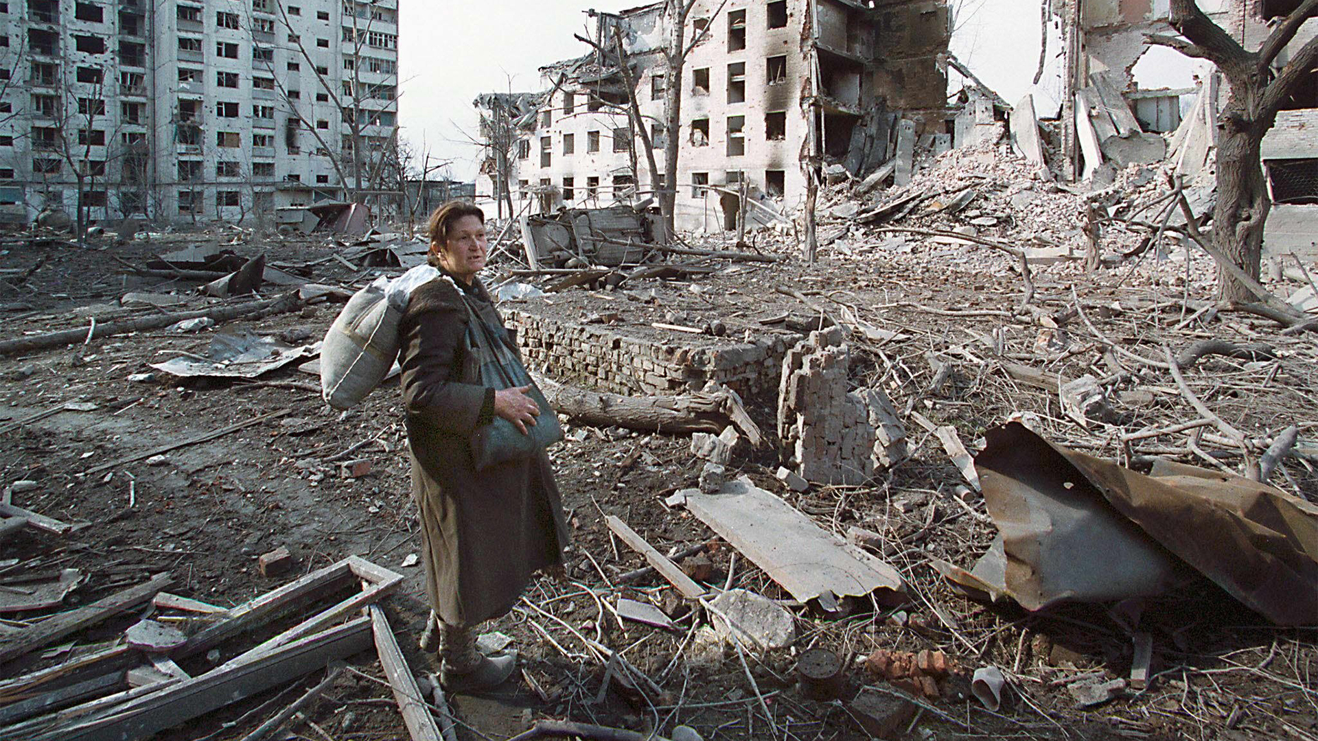 Eine Frau steht mit einer Tasche vor zerstörten Häusern in Grosny (Archivbild vom 23.2.2000). | picture-alliance / dpa