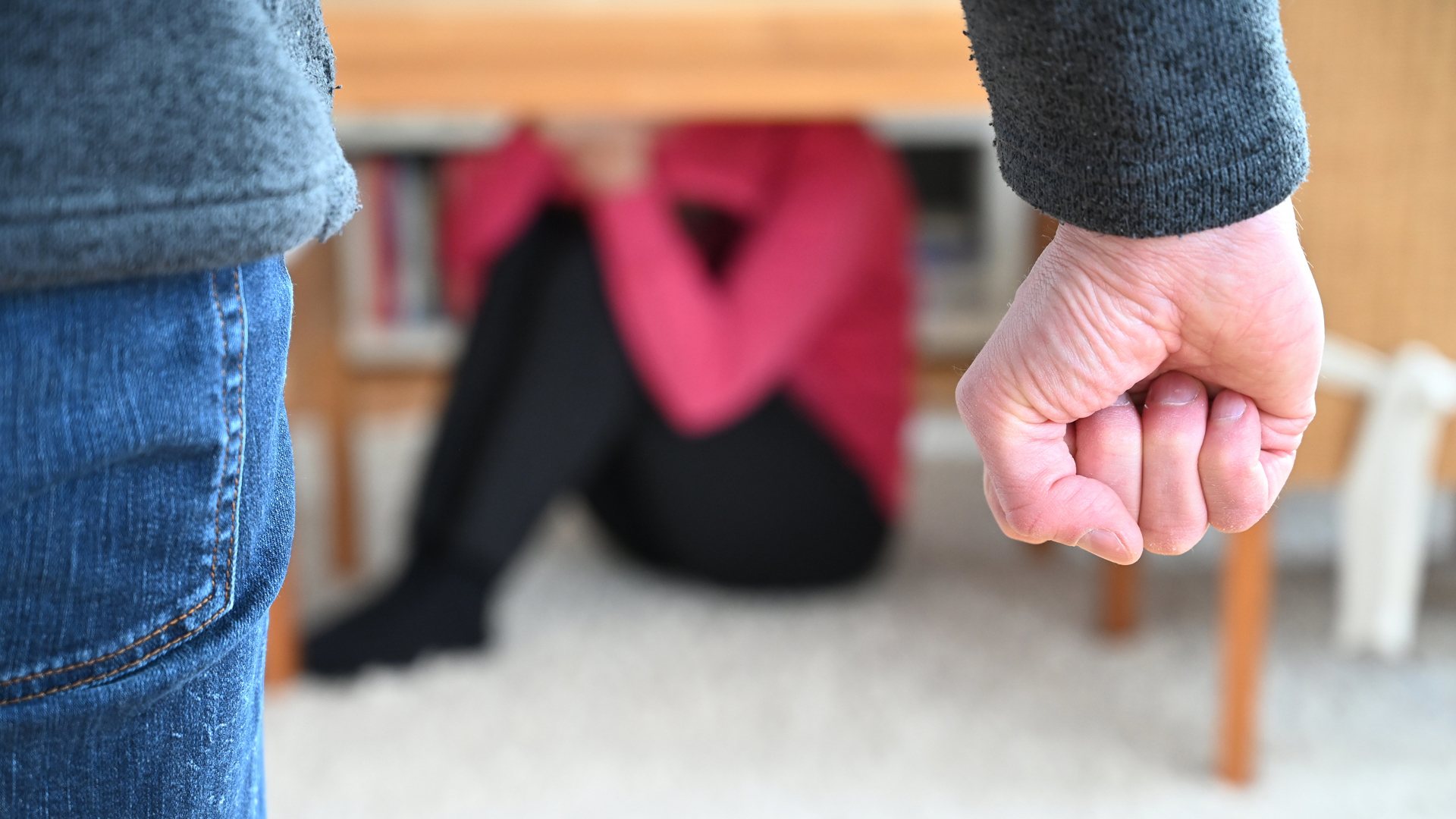 Gestellte Szene zur häuslichen Gewalt: Mann ballt eine Faust, eine Frau sitzt im Hintergrund Schutz suchend unter einem Tisch.