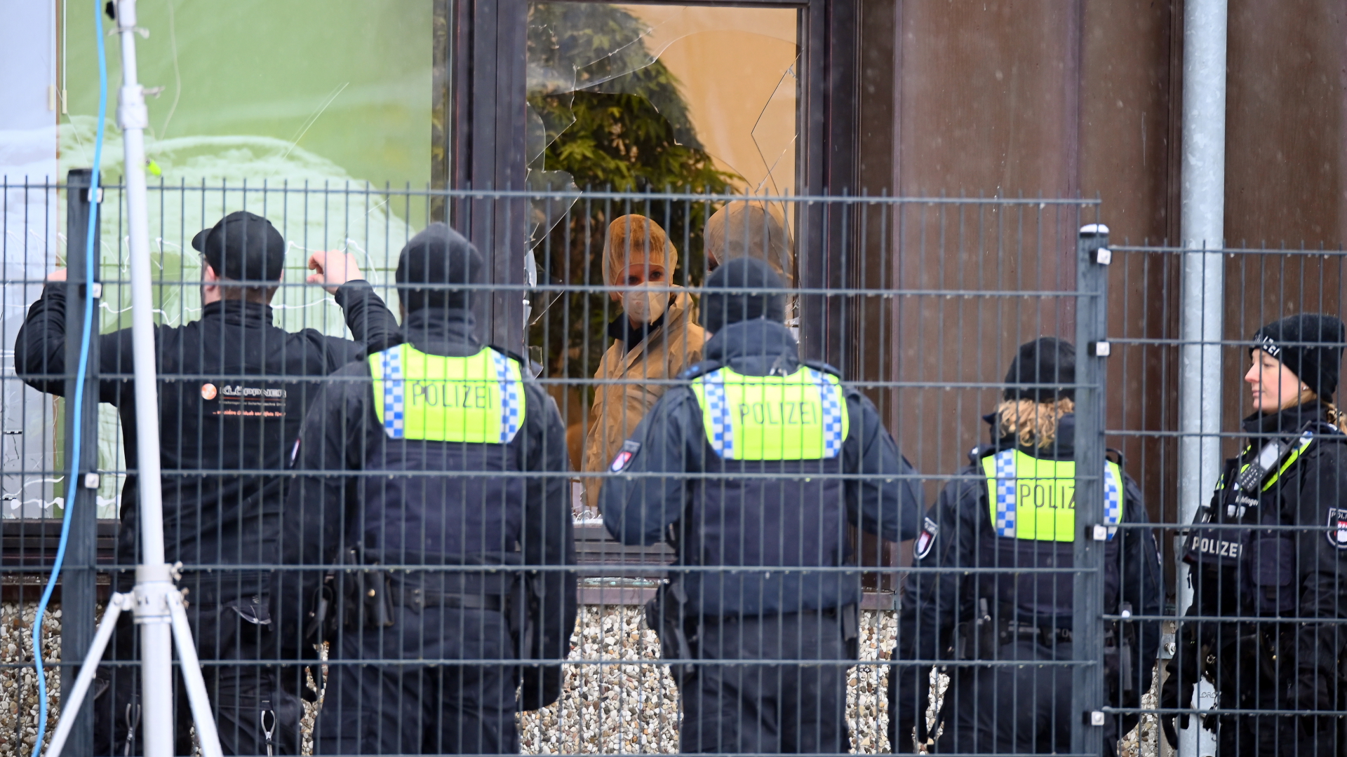 Polizisten sichern in Hamburg-Alsterdorf ein Gebäude, durch eine zerborstene Scheibe ist eine Beamtin in weißem Schutzanzug zu sehen. | dpa