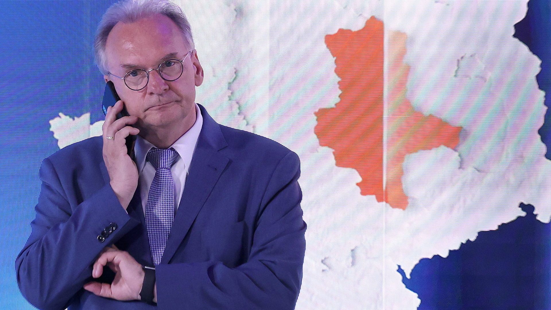 Reiner Haseloff telefoniert vor einem Bildschirm, der die Umrisse Sachsen-Anhalts zeigt.
