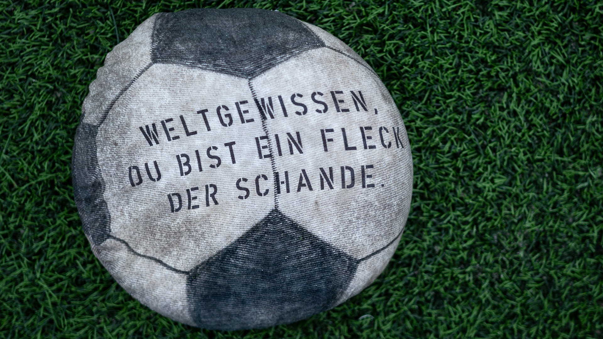 Ein mit Sand gefüllter Stoff-Fußball, auf dem "Weltgewissen, du bist ein Fleck der Schande" steht, liegt auf dem Rasen eines Fußballstadions am Schloss Strünkede in Herne (Nordrhein-Westfalen).  | dpa