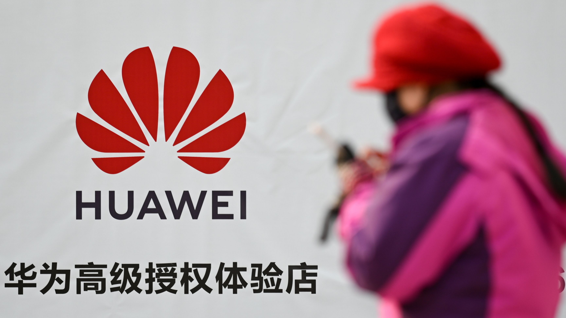 Das Logo von Huawei an einer weißen Hauswand, an der schemenhaft eine Frau vorbeiläuft, die auf ihr Handy blickt. | AFP
