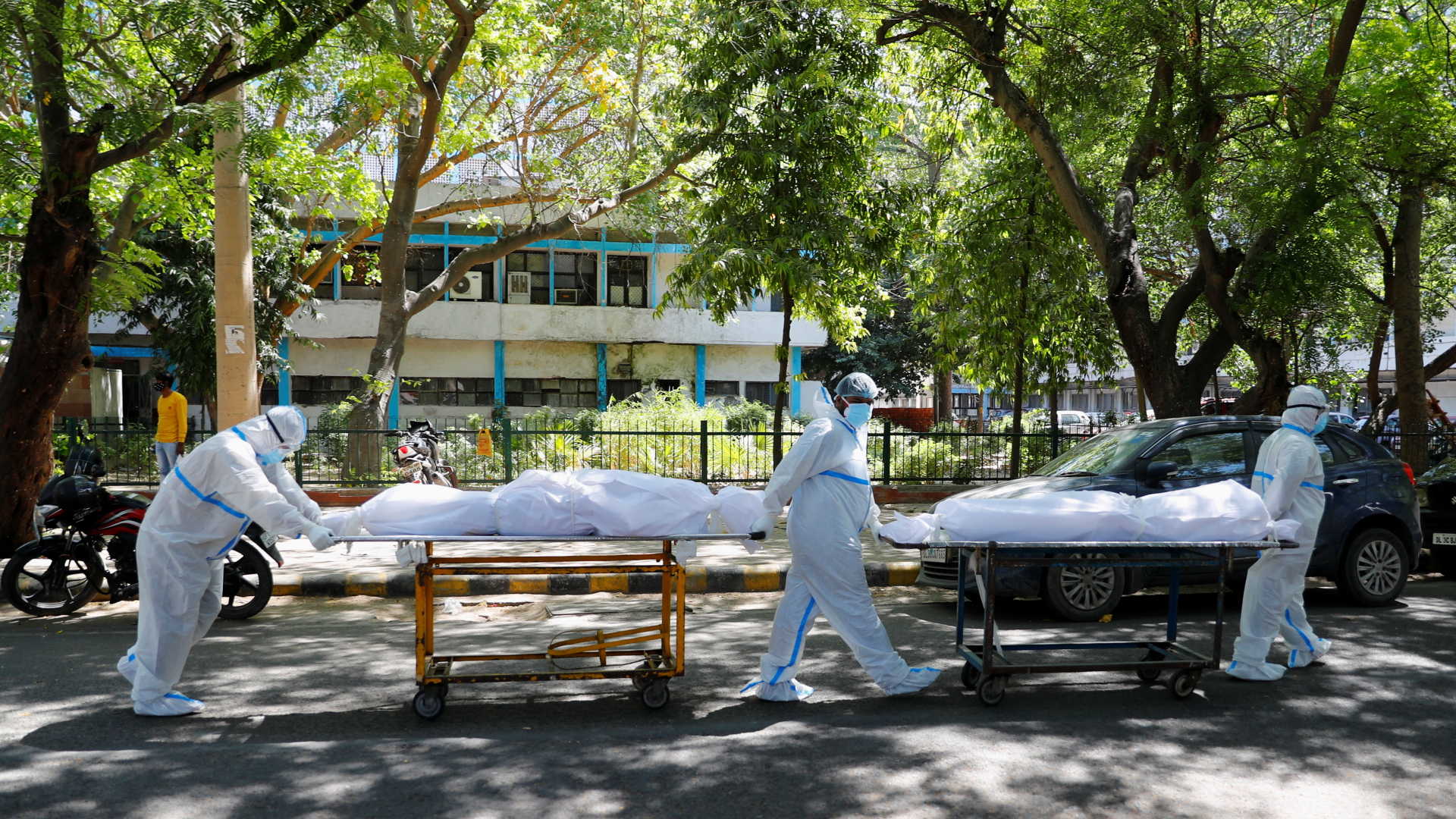 In Neu-Delhi transportieren drei Männer in Schutzkleidung zwei Ambulanzliegen mit zwei in weißen Stoff eingehüllten Corona-Toten. | REUTERS