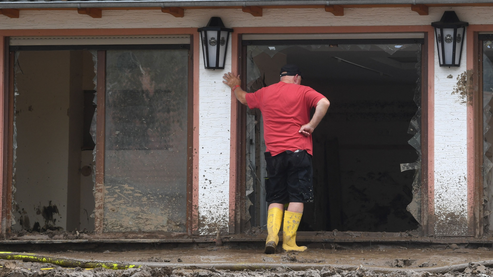 Ein Mann in Gummistiefeln steht im Schlamm und schaut in ein zerstörtes Haus hinein. | AFP