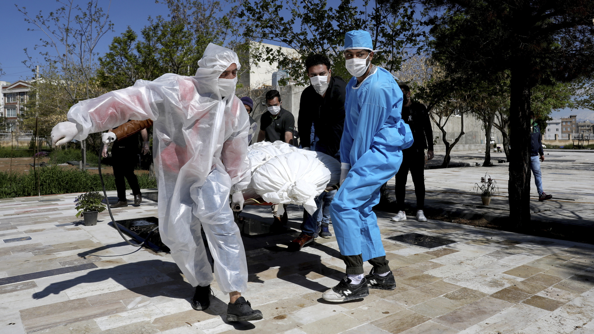 Männer in Schutzkleidung tragen einen in weißes Tuch gehüllten Leichnam eines Corona-Toten zu einem Friedhof in Teheran. | dpa
