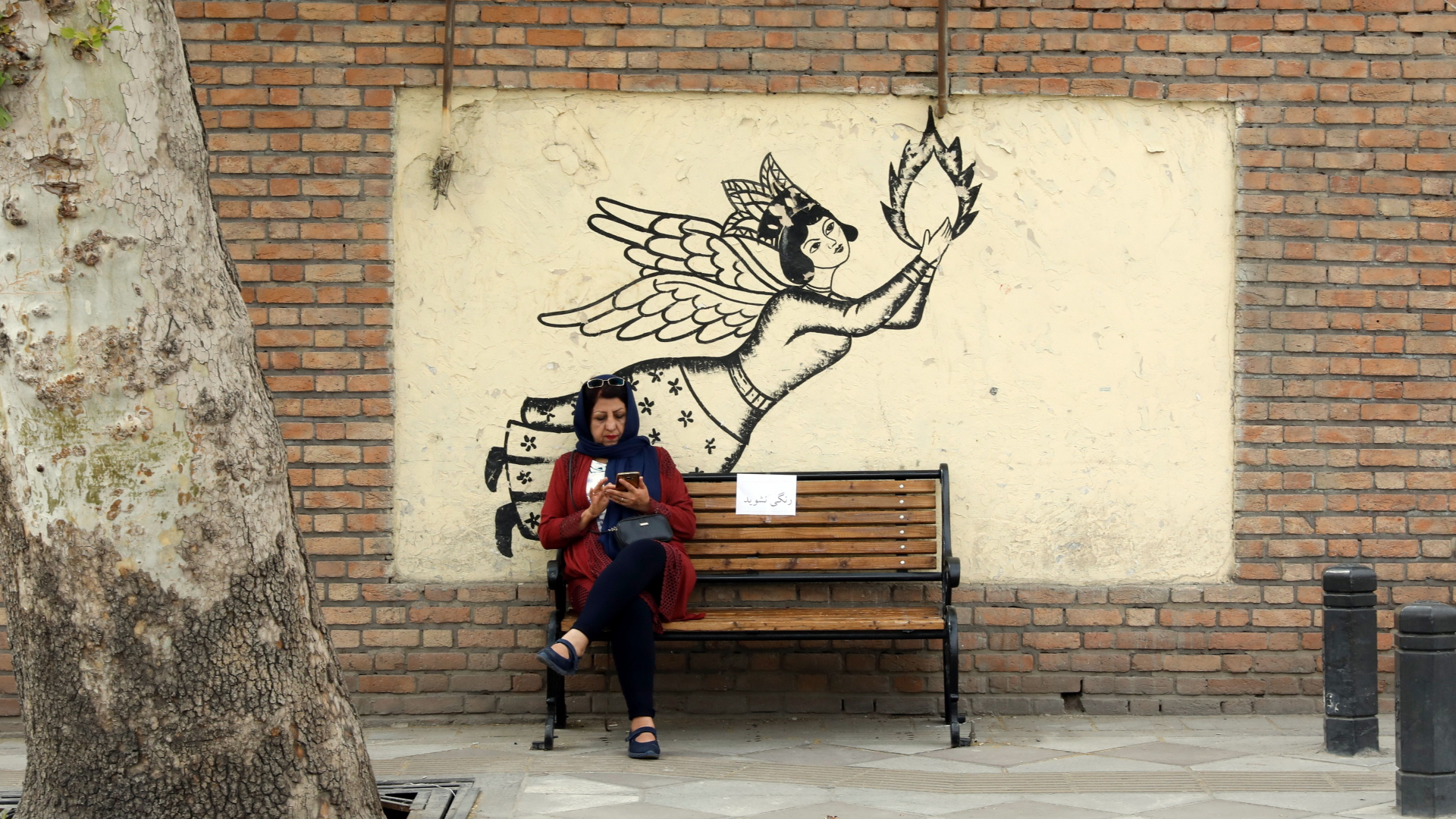 In Teheran hat sich eine Frau auf eine Bank gesetzt und blick auf ihr Handy. | ABEDIN TAHERKENAREH/EPA-EFE/REX/