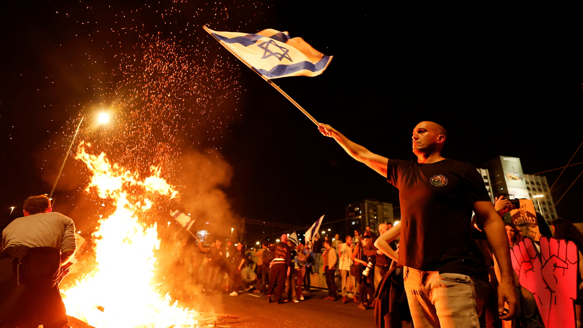 Ein Demonstrant in Tel Aviv schwenkt eine israelische Fahne, neben ihm brennt ein Feuer auf einer Straße | REUTERS