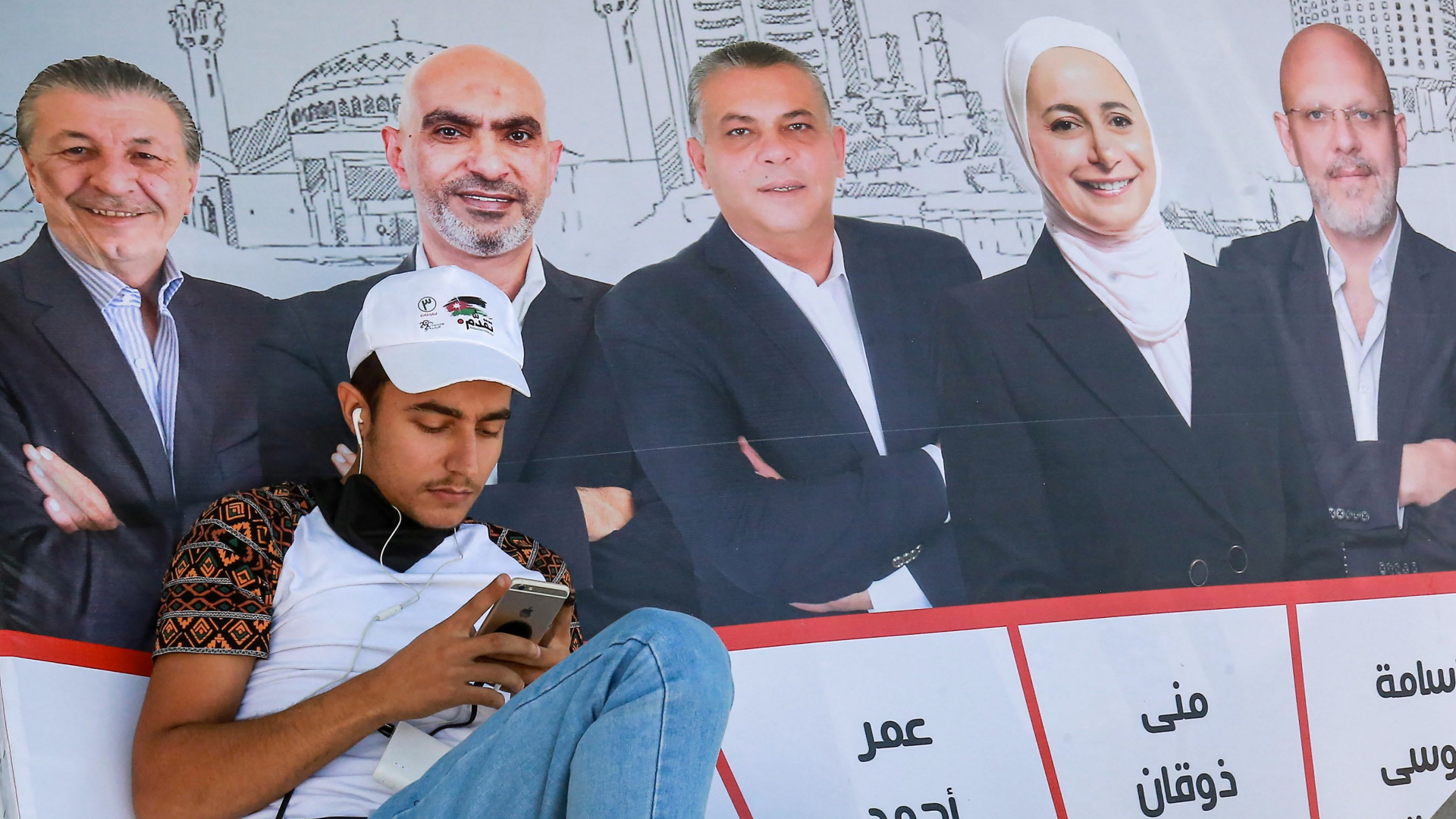 Ein junger Mann sitzt in Amman vor Wahlplakaten und schaut auf sein Handy. | AFP