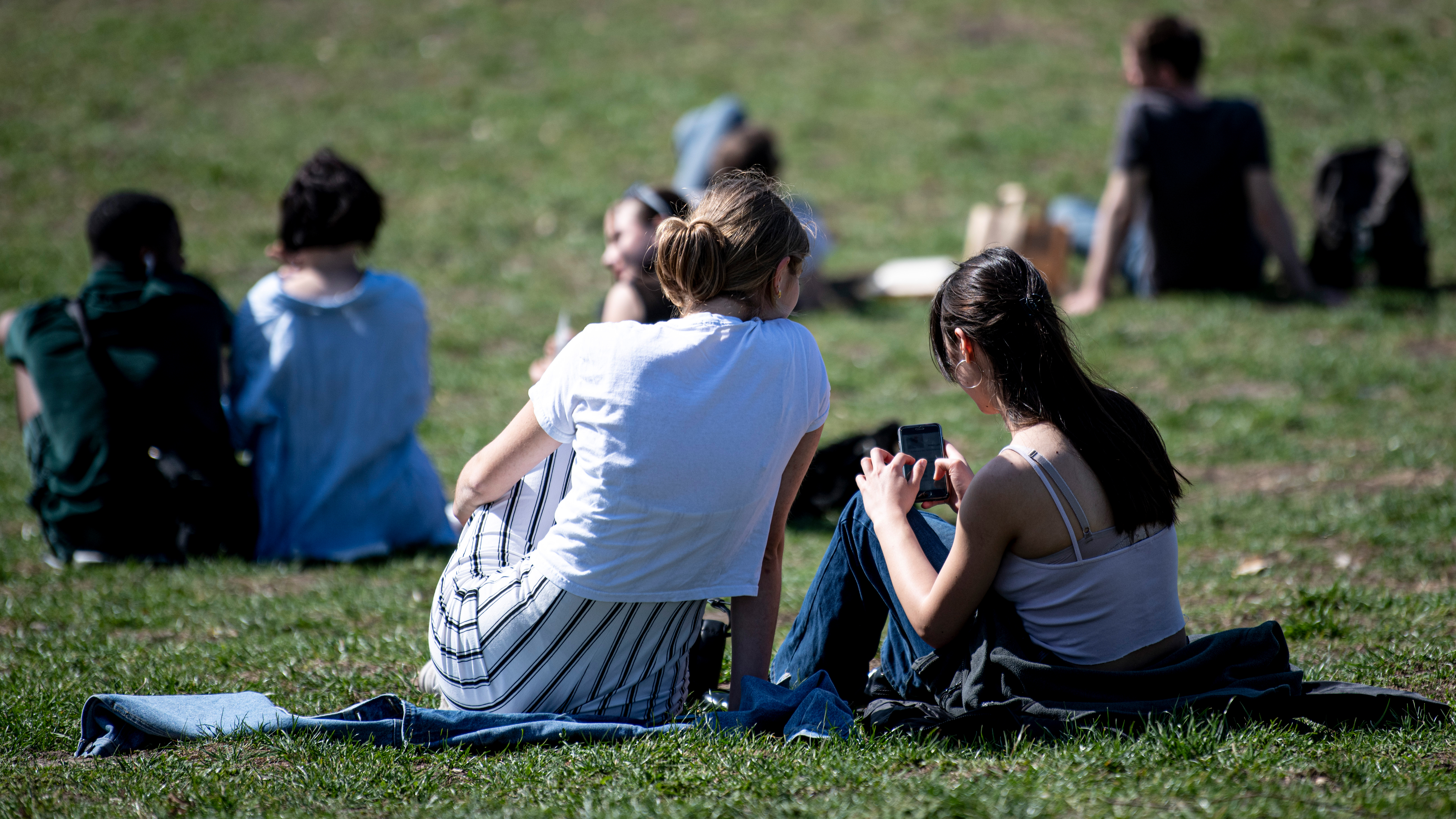 Zwei junge Frauen sitzen in einem Park und schauen auf ein Smartphone. | picture alliance/dpa