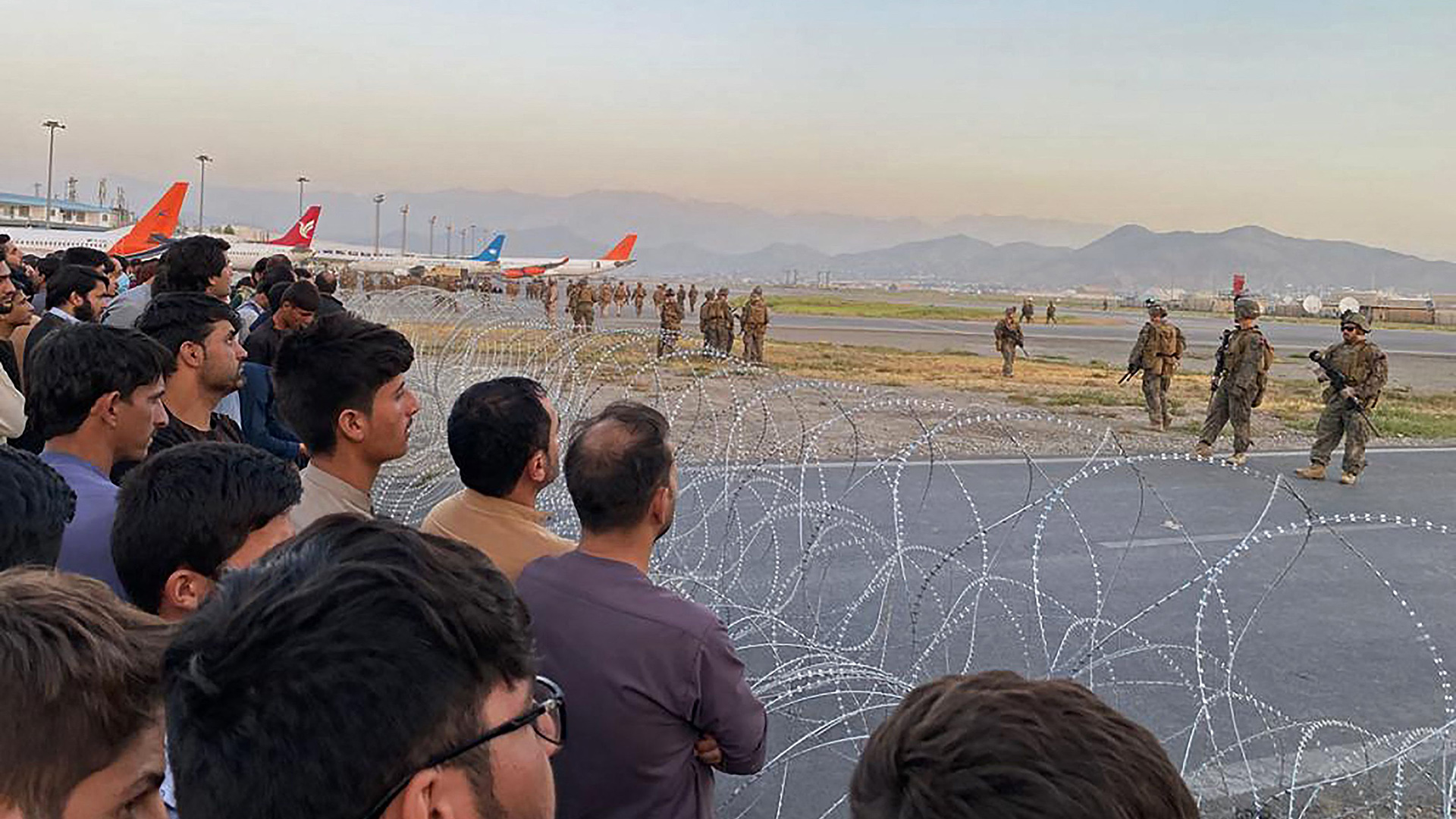 Menschen stehen am Stacheldraht des Flughafens in Kabul, auf dem Militär zwischen Flugzeugen steht. | AFP