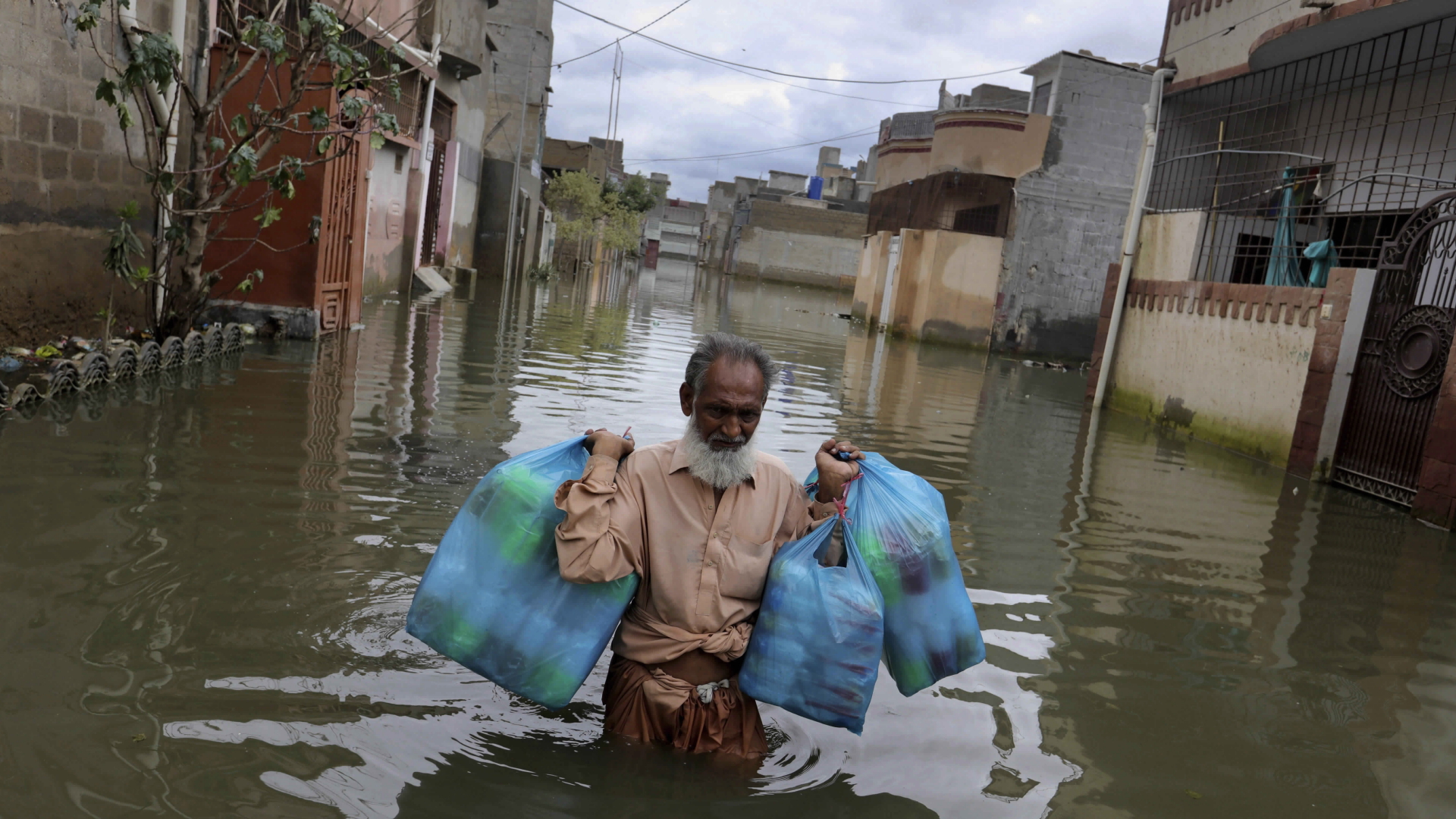 Ein Mann steht bis zur Hüfte im Wasser auf einer Straße im pakistanischen Karatschi und trägt gefüllte Plastiktüten. | dpa