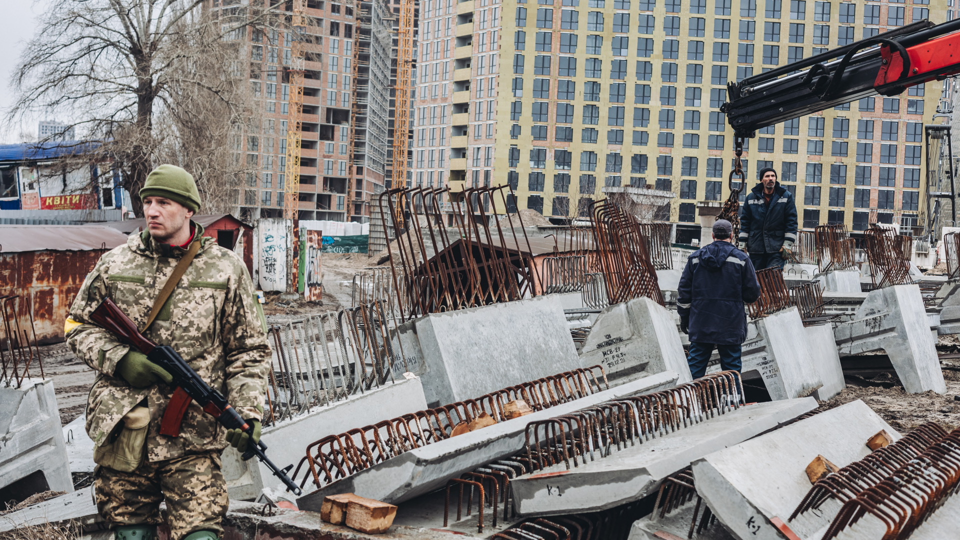Arbeiter laden in Kiew Betonplattenab, während ein bewaffneter ukrainischer Mann Wache steht.  | dpa