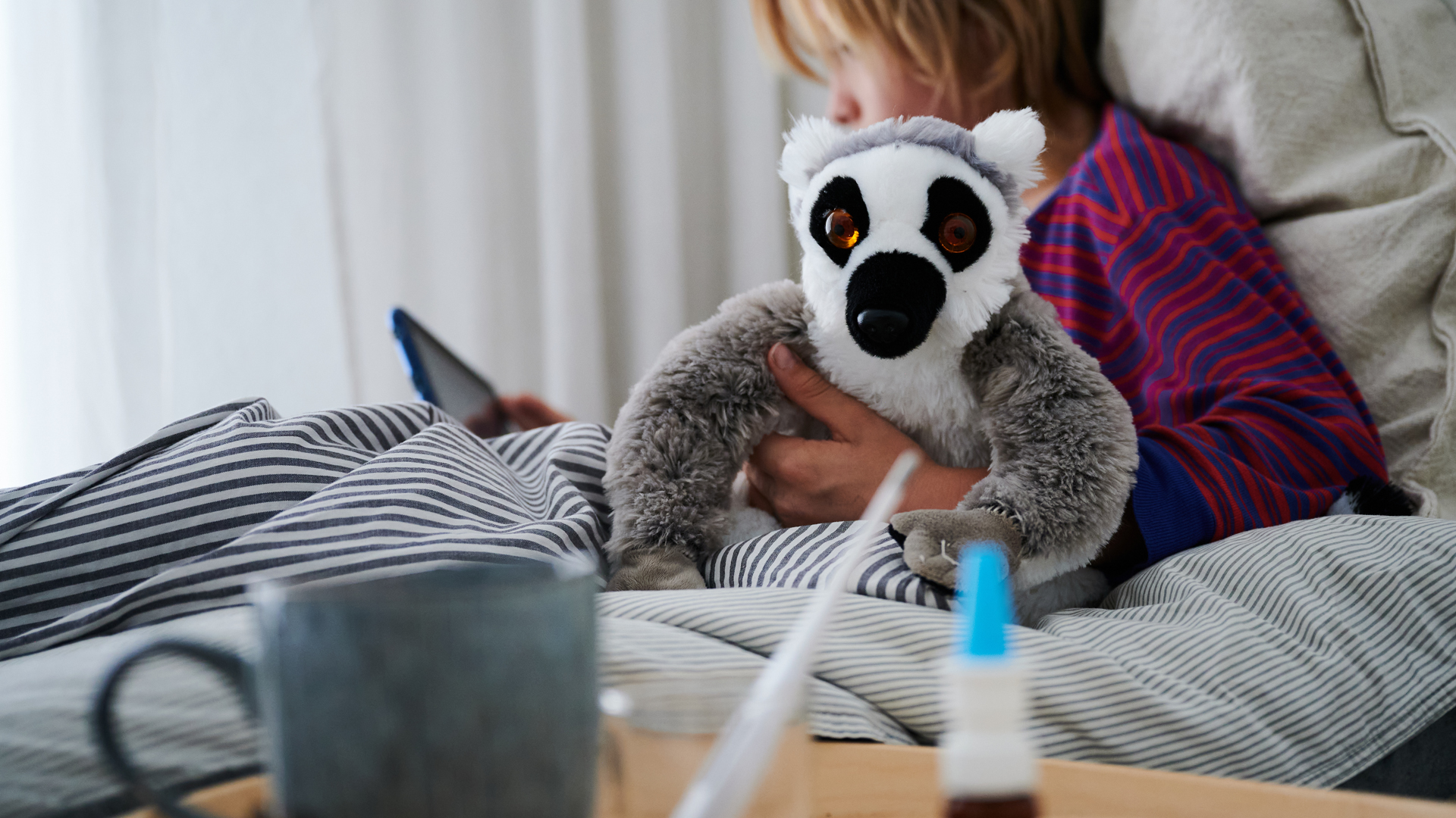Ein Fieberthermometer, Nasenspray und eine Tasse Tee stehen vor einem Bett, in dem ein Kind mit Kuscheltier sitzt und auf ein Tablet schaut. | picture alliance/dpa