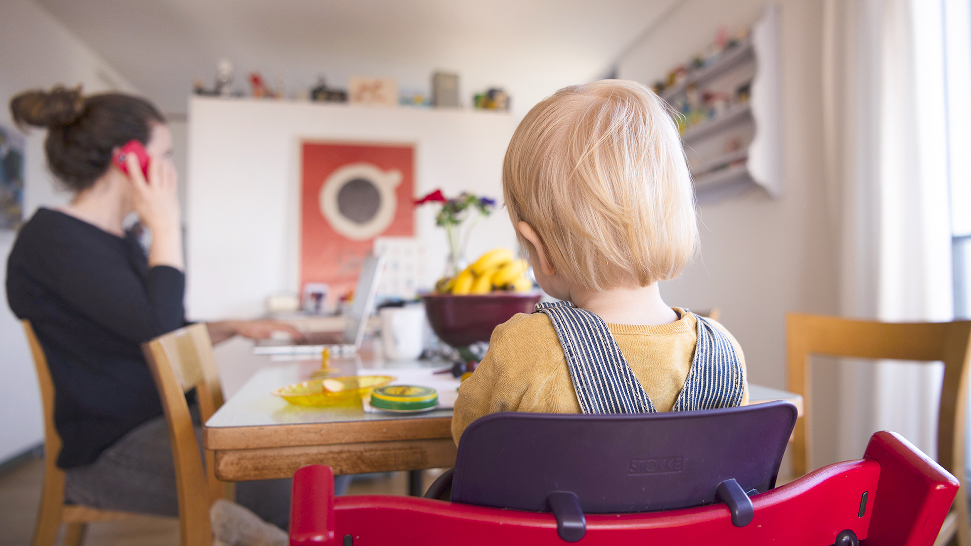  Eine Frau sitzt im Homeoffice an ihrem Laptop und telefoniert, während ihr Kind neben ihr in einem Kinderstuhl am Tisch sitzt.