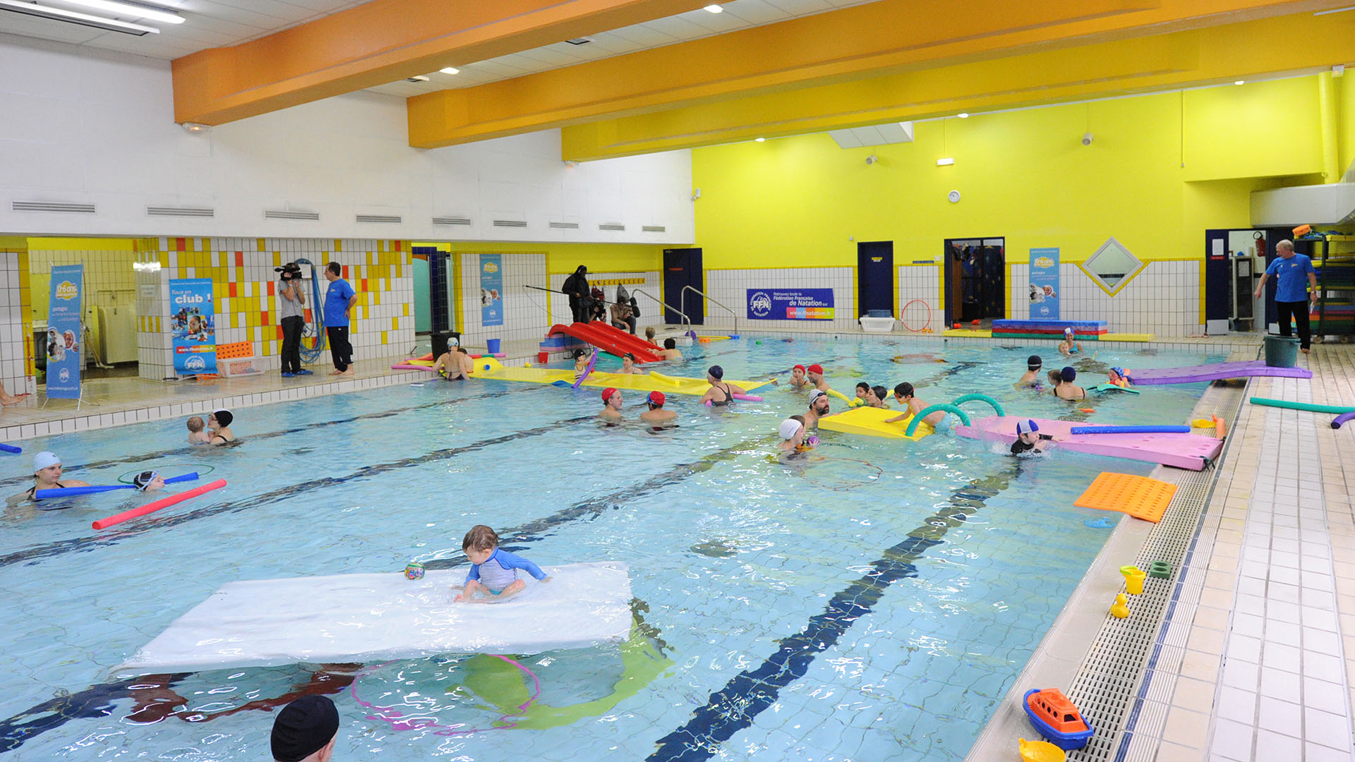 Kinder und Erwachsene mit verschiedenen Schwimmhilfen in einer Schwimmhalle.
