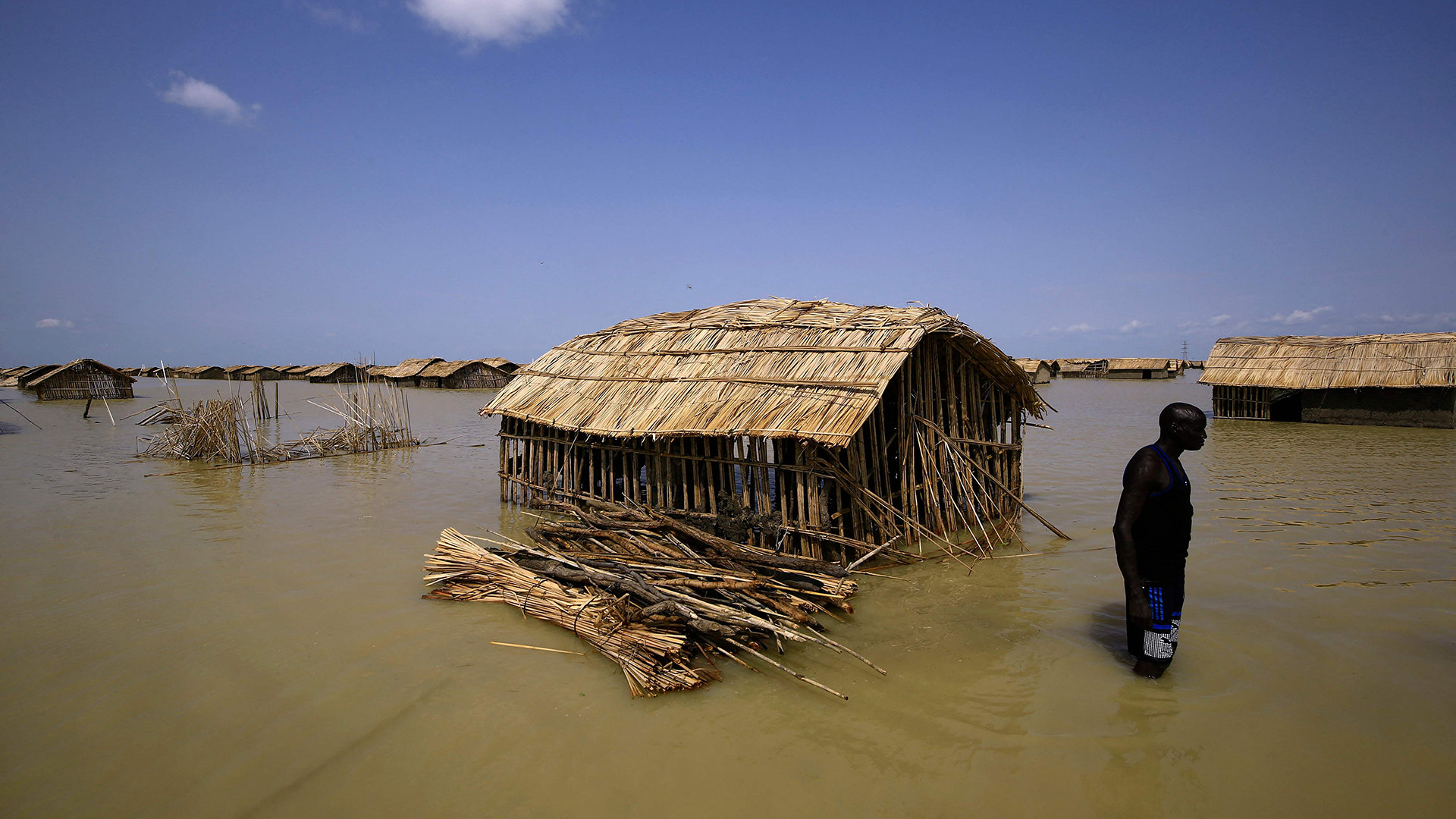 Ein Mann im Sudan watet durch hohes Wasser zwischen Hütten, die unter Wasser stehen.