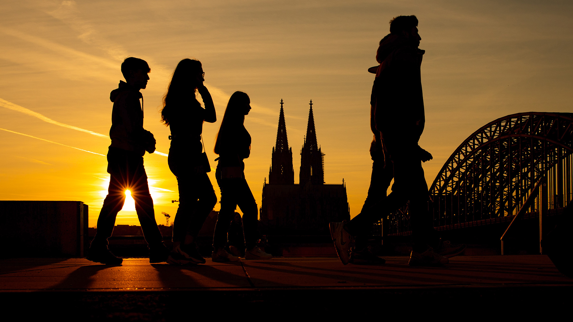 Jugendliche laufen bei untergehender Sonne über die Rheinpromenade. Im Hintergrund ist die Silhouette des Kölner Doms zu sehen. | dpa