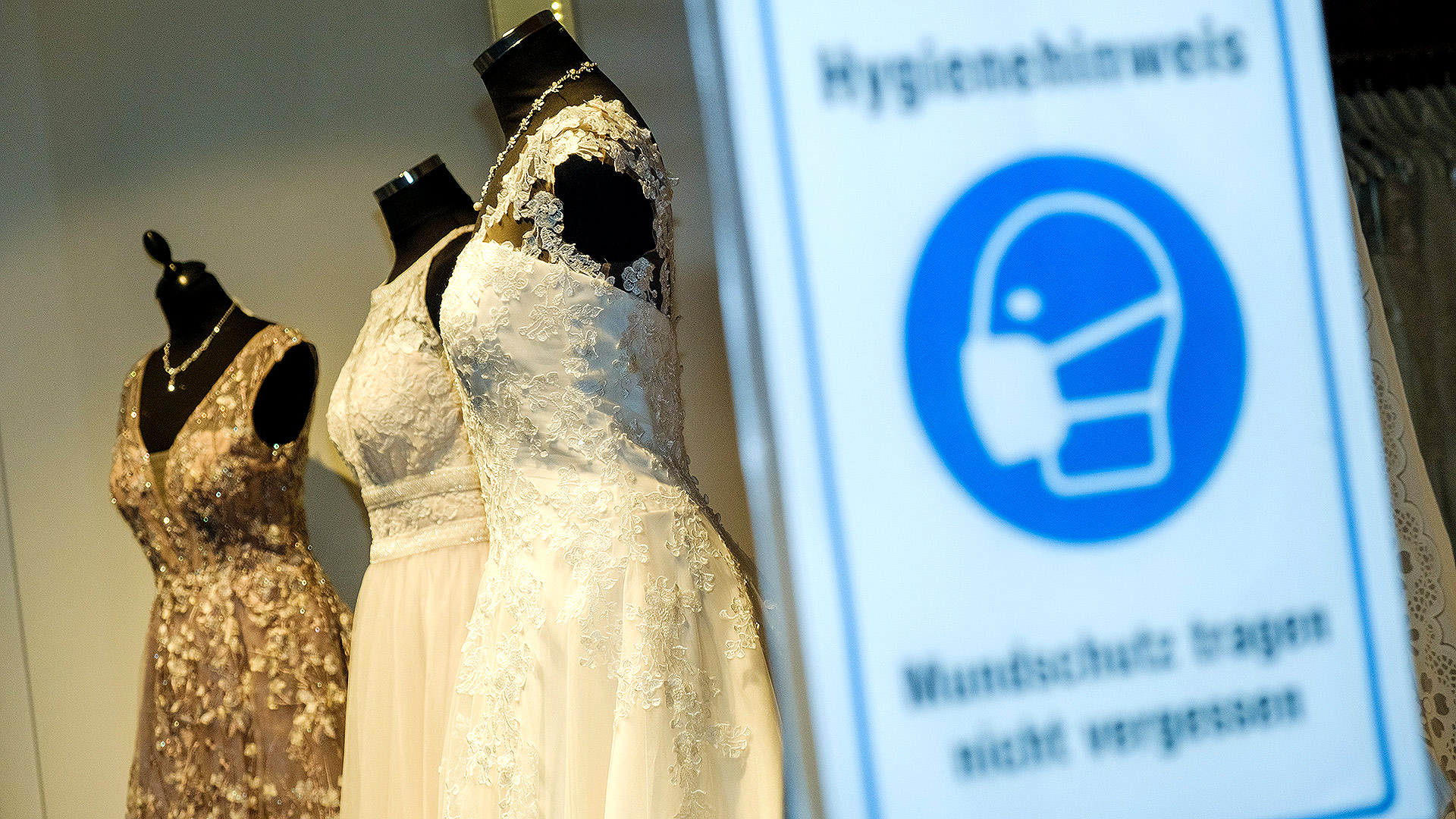 Brautkleider in einem Schaufenster und im Vordergrund ein Hinweisschild zur Maskenpflicht während der Corona-Pandemie | 