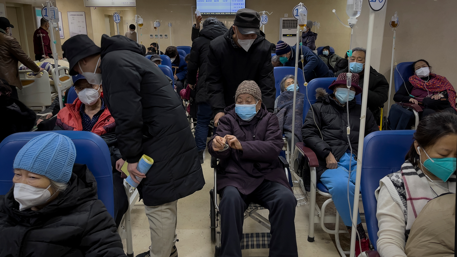 Ein Mann schiebt eine ältere Frau im Rollstuhl an Patienten vorbei, die in einem Korridor der Notaufnahme eines Krankenhauses intravenöse Infusionen erhalten. | dpa