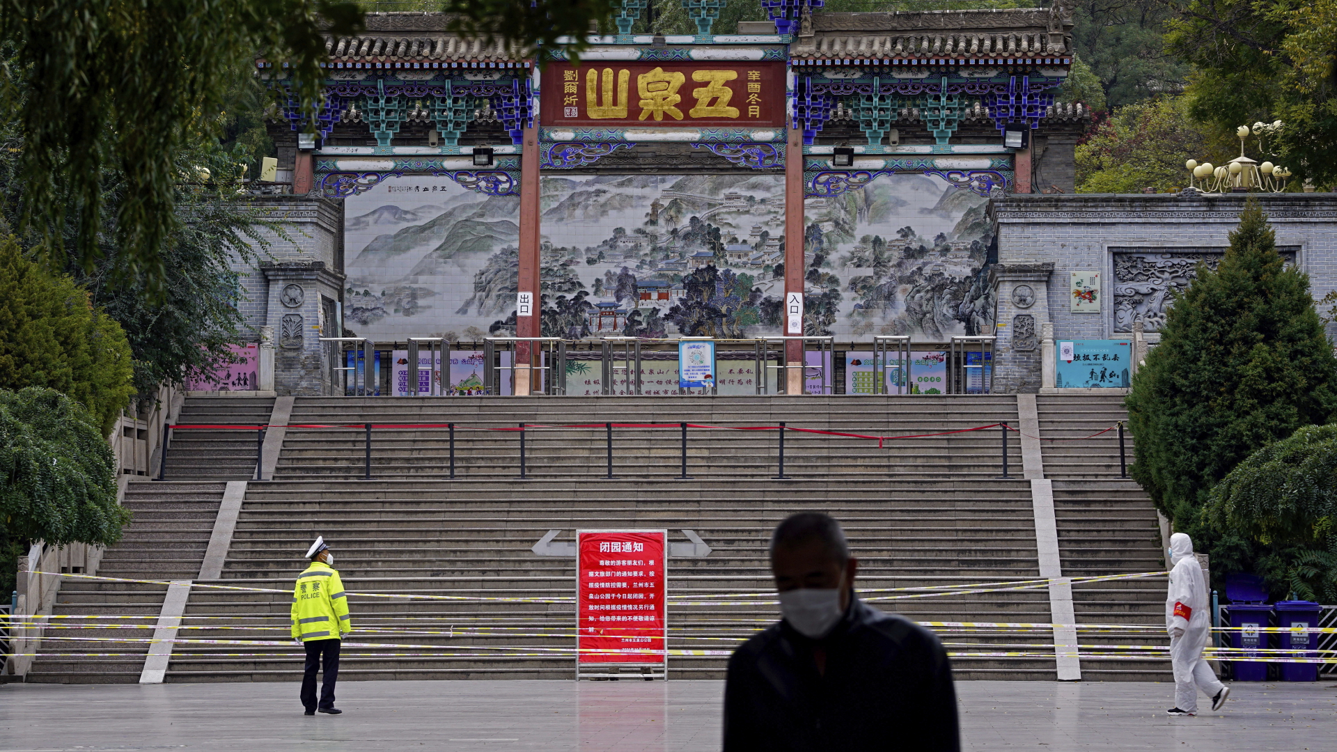 Ein Polizist (l) trägt gelbe Warnkleidung und einen Mund-Nasen-Schutz, als er vor einer Absperrung steht, die am Eingang zum Wuquan-Berg befestigt ist. | dpa