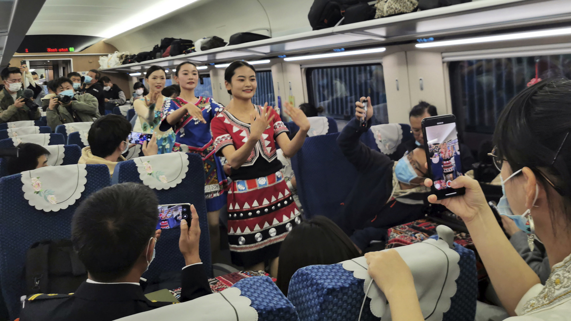 Fahrgäste zücken ihre Handys, als eine Tänzerinnengruppe in Tracht durch den Mittelgang geht. | AP
