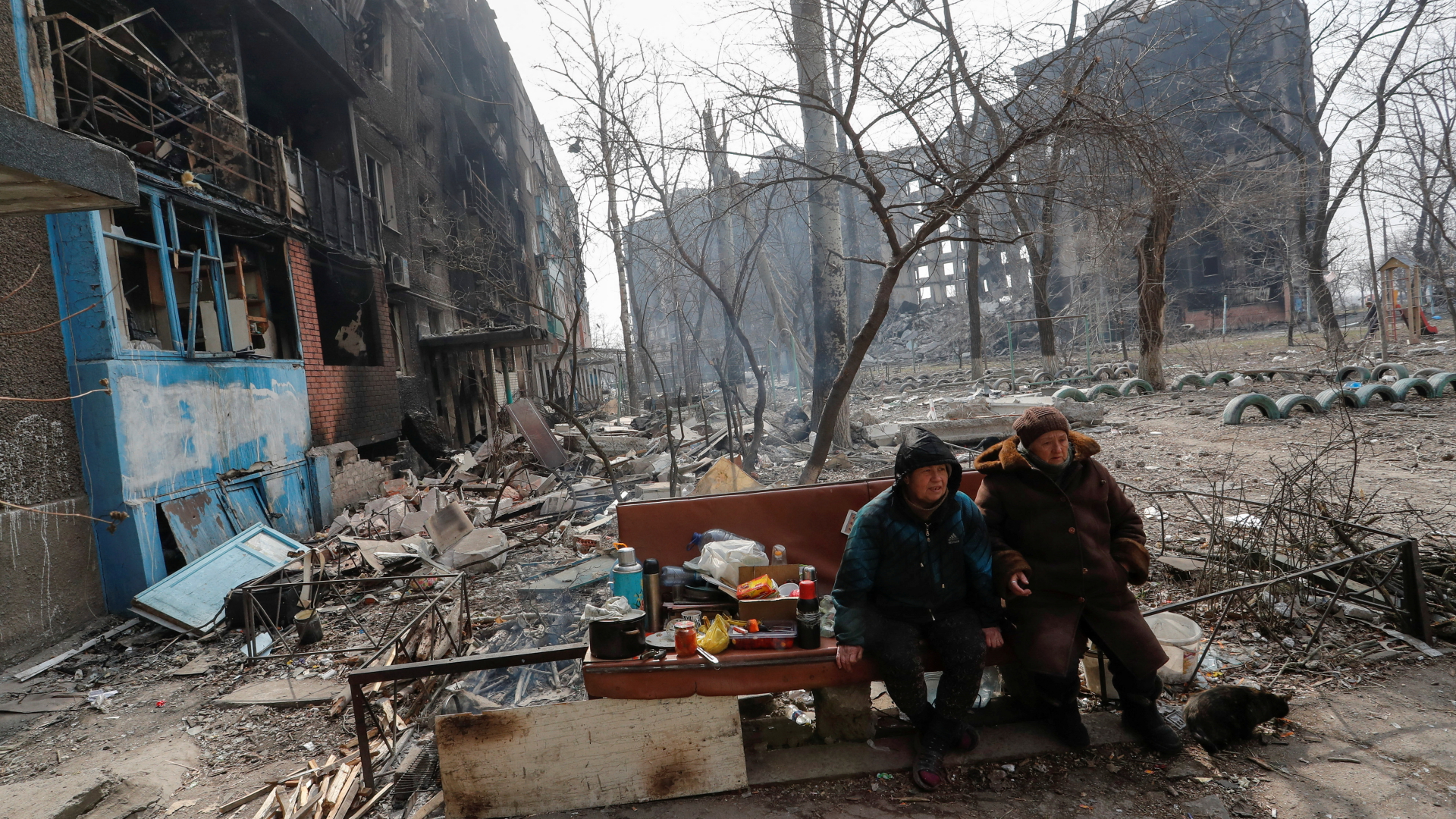 Zwei Frauen sitzen auf einer Bank in Mariupol, im Hintergrund kaputte Gebäude.