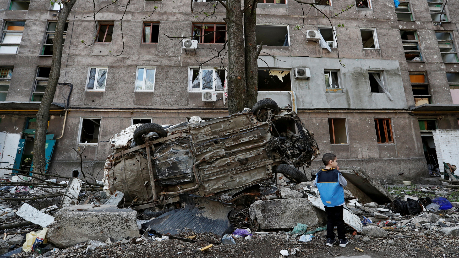Ein Junge steht neben einem Autowrack vor einem Wohnhaus, das während des Ukraine-Russland-Konflikts in der südlichen Hafenstadt Mariupol beschädigt wurde | REUTERS