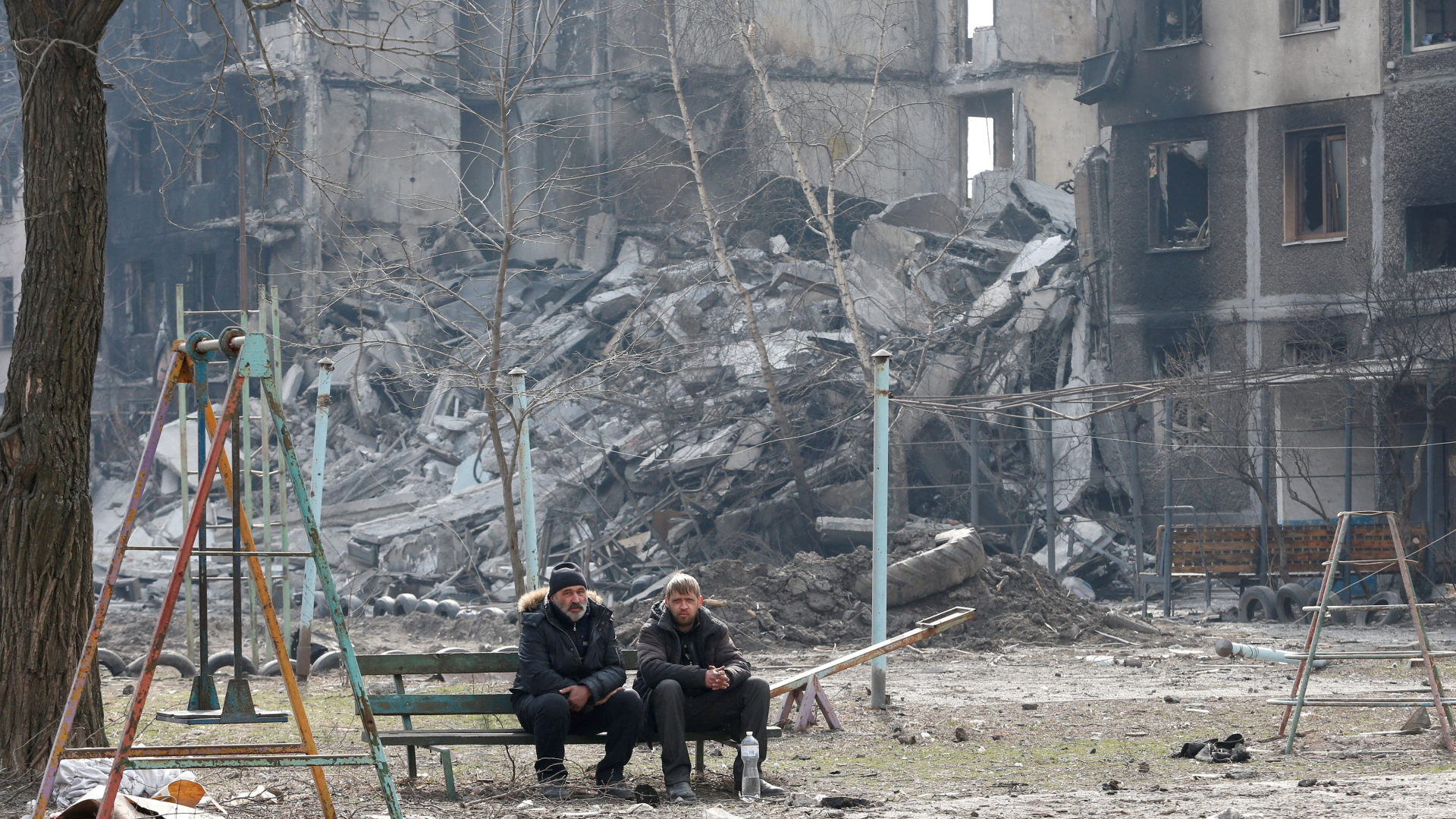 Zwei Männer sitzen auf einer Spielplatz-Bank vor zerstörten Häusern in Mariupol. | REUTERS