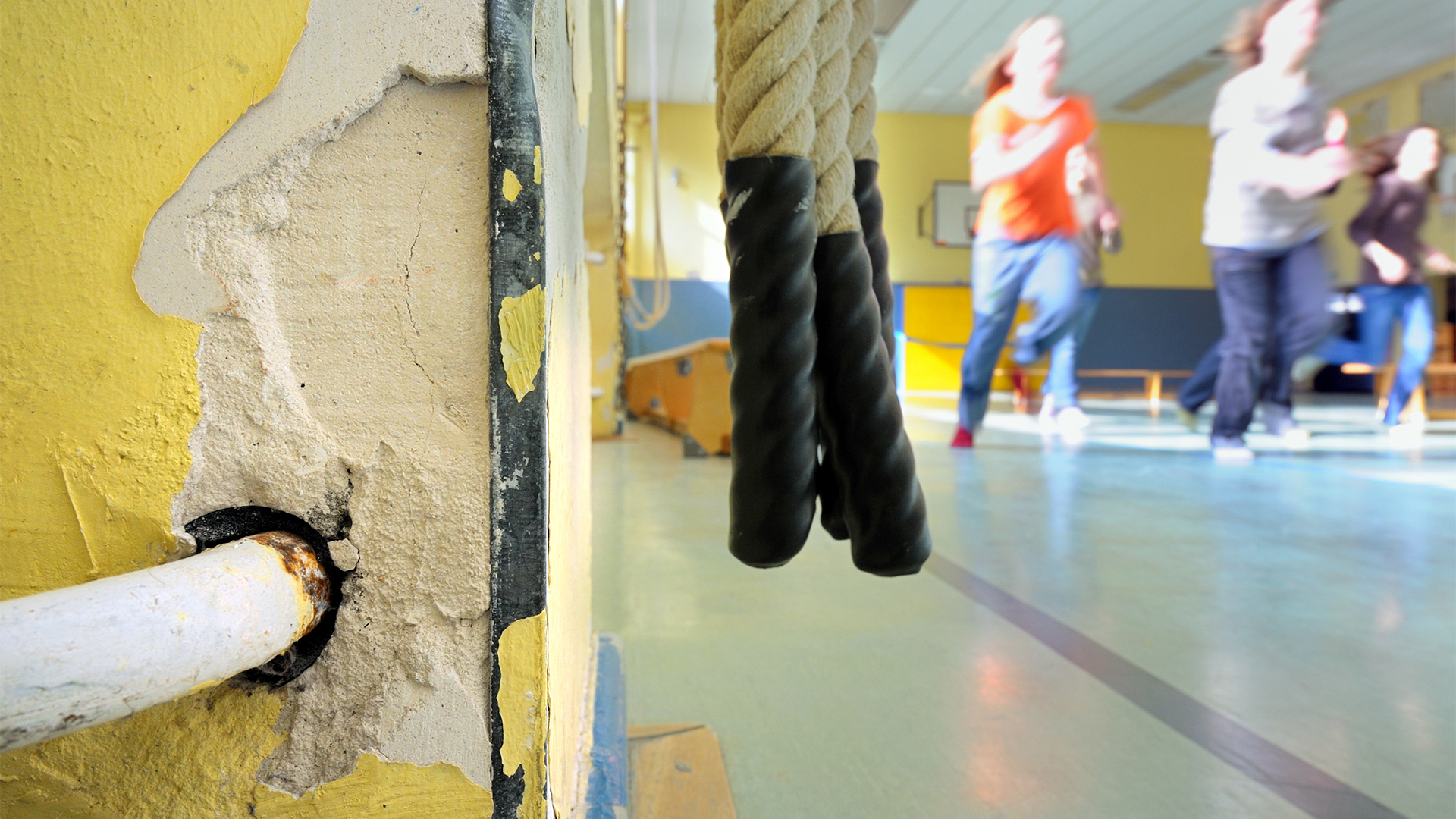 Kinder laufen in der Turnhalle einer Schule in Darmstadt an der Zuleitung eines Heizkörpers vorbei, in dessen Umgebung bereits der Putz abgebröckelt ist. | picture alliance / Uwe Anspach/d