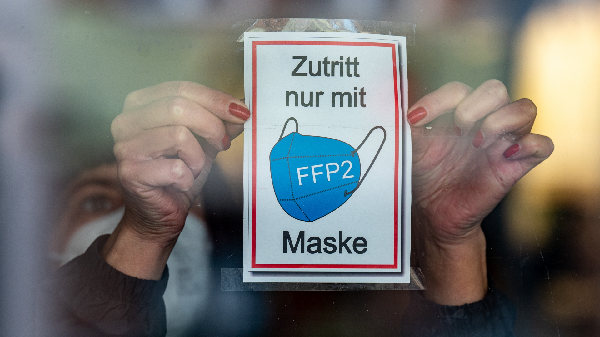 Hände kleben Schild mit dem Tragen einer FFP2-Maske als Zugangsvoraussetzung an eine Glastür | dpa