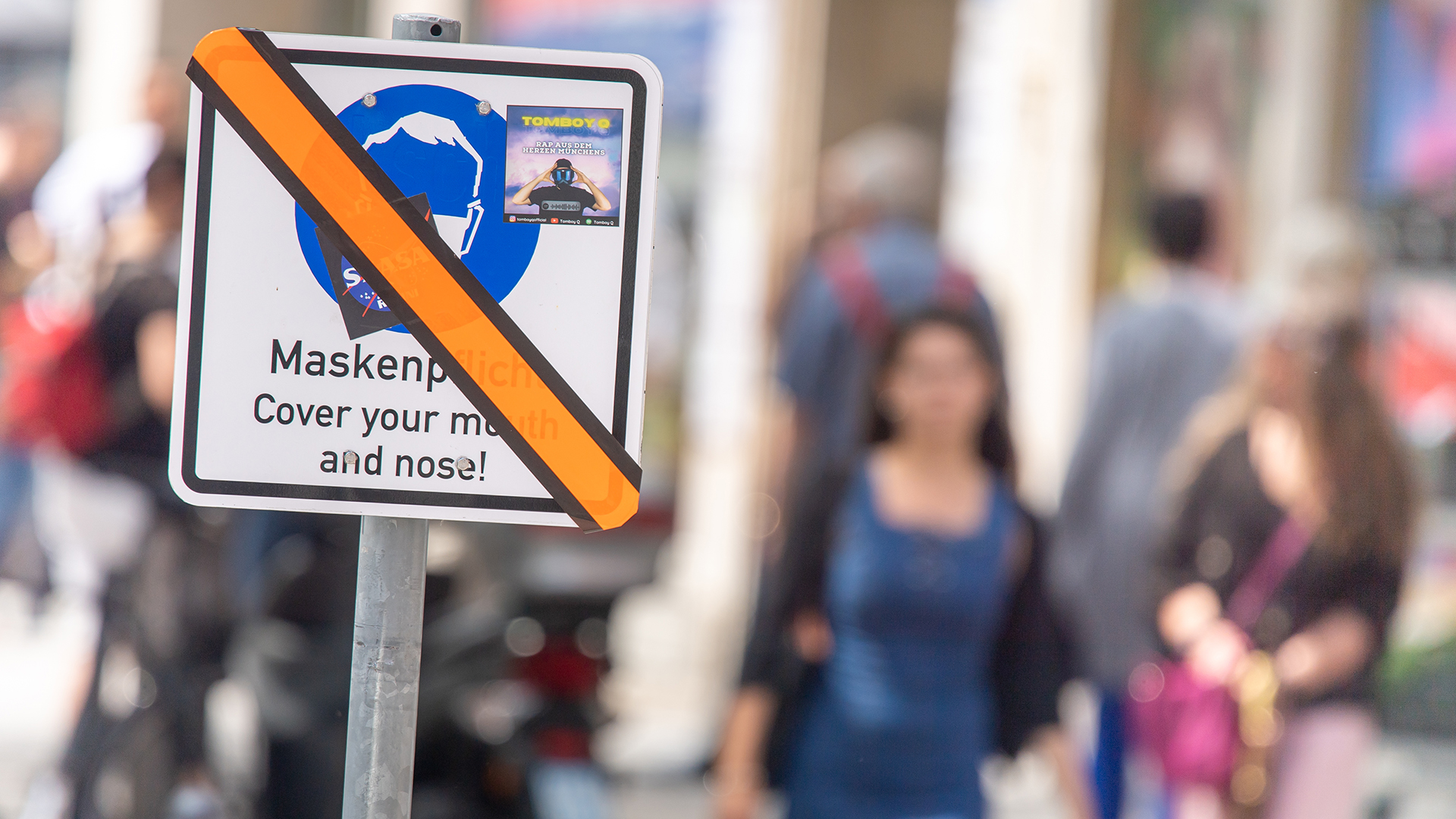 Über ein Schild mit dem Hinweis zur Maskenpflicht in München wurde mit einem orangefarbenen Streifen überklebt