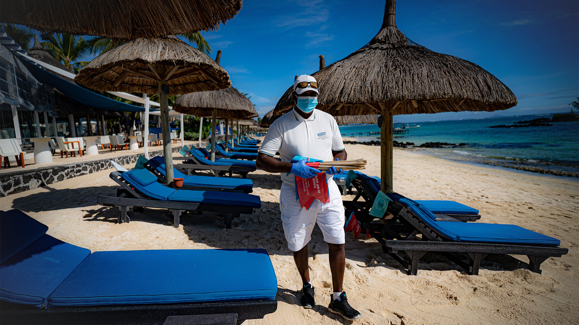 Ein Hotelangestellter auf Mauritius mit medizinischem Mund-Nasen-Schutz und Handschuhen desinfiziert Strandliegen am Strand. | picture alliance/dpa/PA Wire