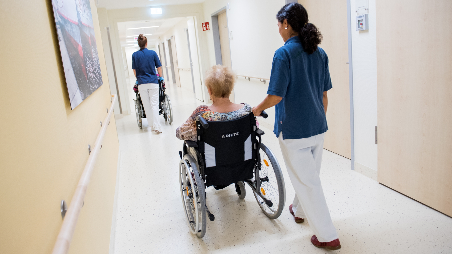 Zwei Pflegerinnen schieben Patienten in Rollstühlen über den Flur eines Krankenhauses.  | dpa