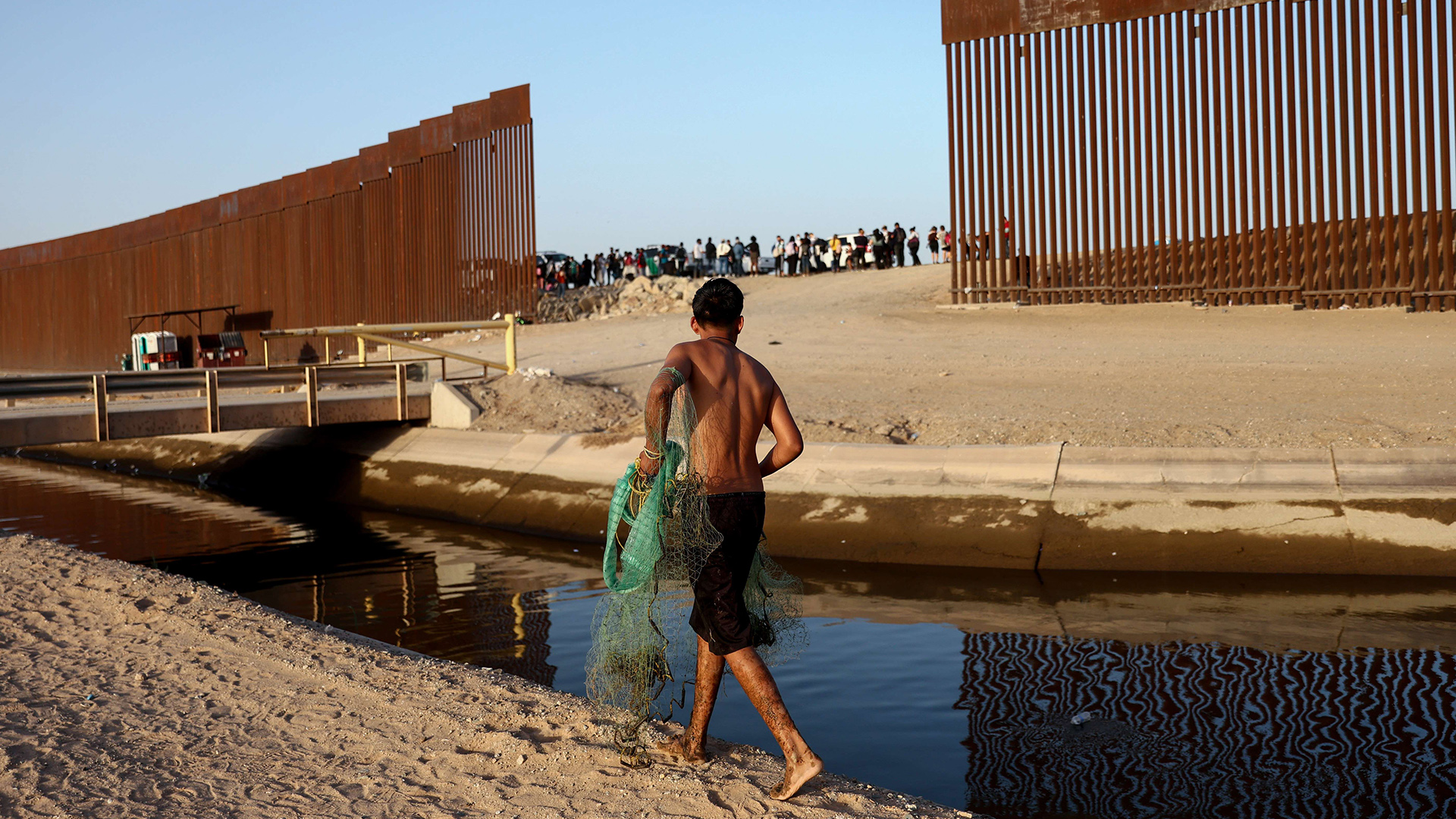 Ein Mann mit Fischernetzt blickt auf Menschen, die durch die Lücke im Grenzzaun zu den USA gehen. | AFP