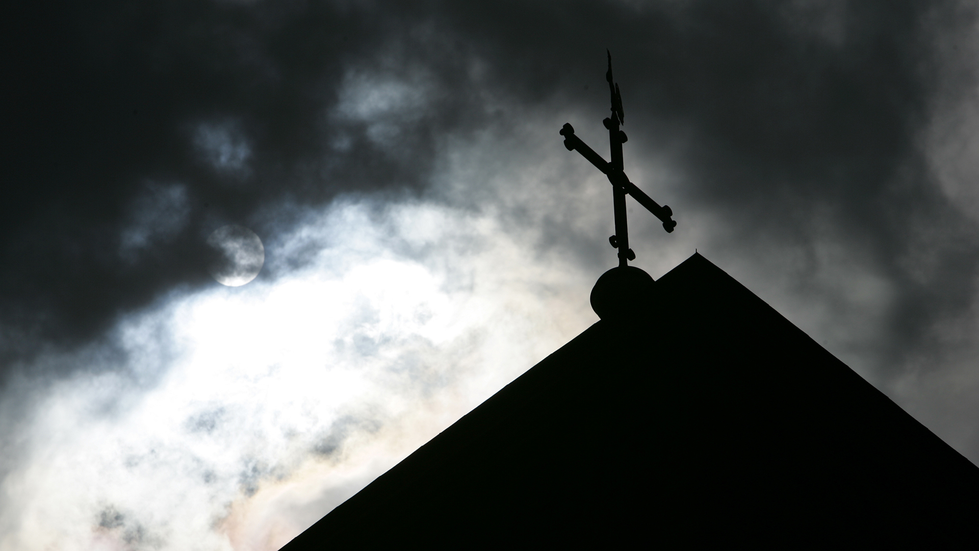 Im Gegenlicht und vor wolkenverhangenem Himmel ist eine Kirchturmspitze mit Kreuz zu sehen