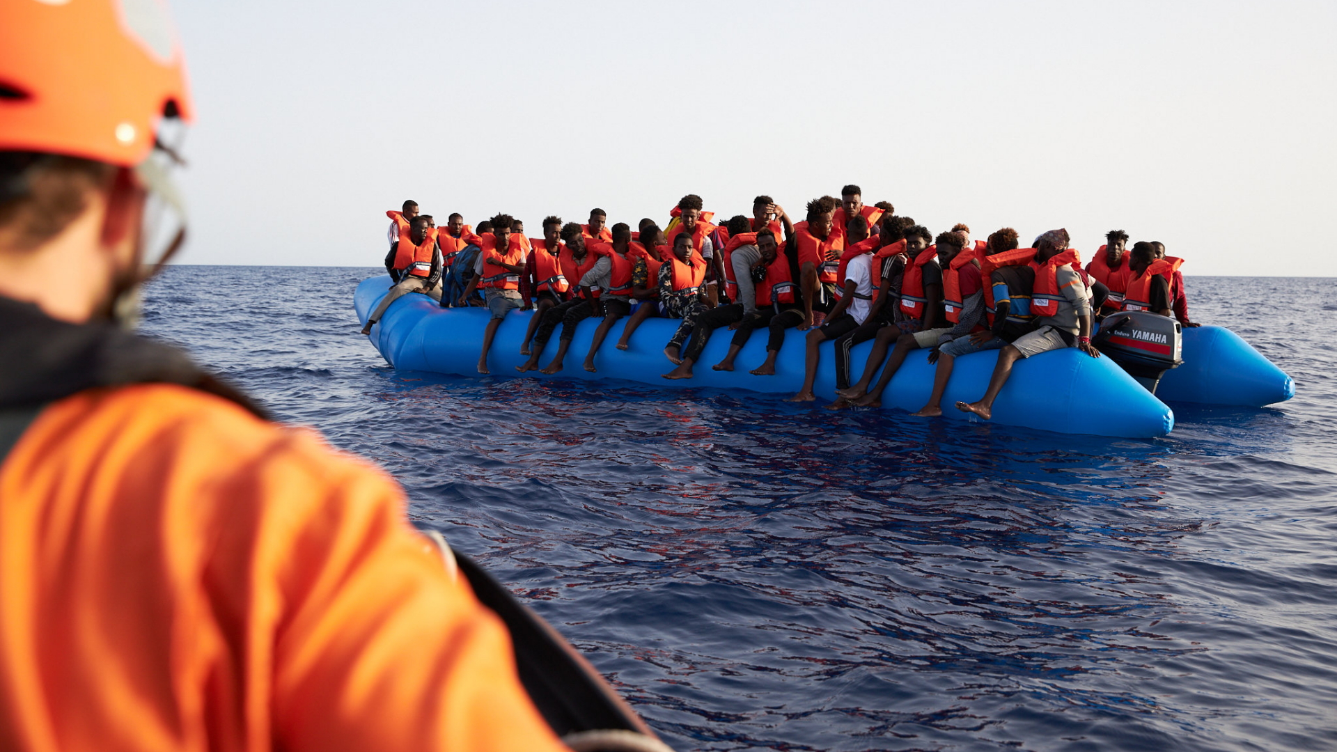 Ein Mitglied der Sea-Eye-Hilfsorganisation blickt auf ein Schlauchboot voller Migranten, das im Mittelmeer treibt. | dpa