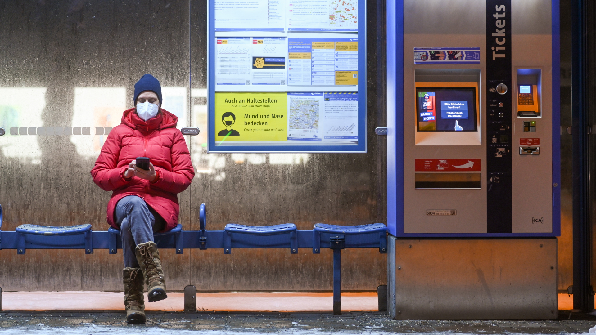 Bayern:  Eine Frau mit Maske sitzt an einer Trambahn-Haltestelle in München. | dpa