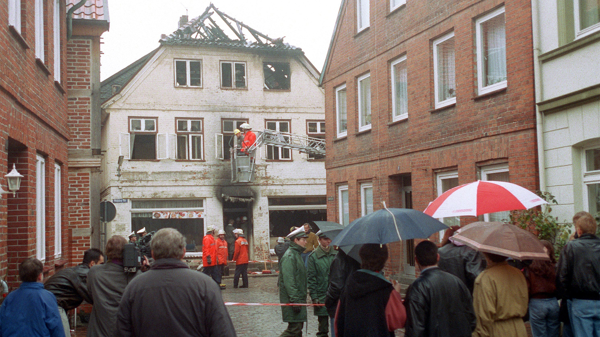 Das Archivbild vom 23.11.1992 zeigt Schaulustige und Feuerwehrleute vor dem Haus in Mölln | picture alliance / Rolf Rick/dpa