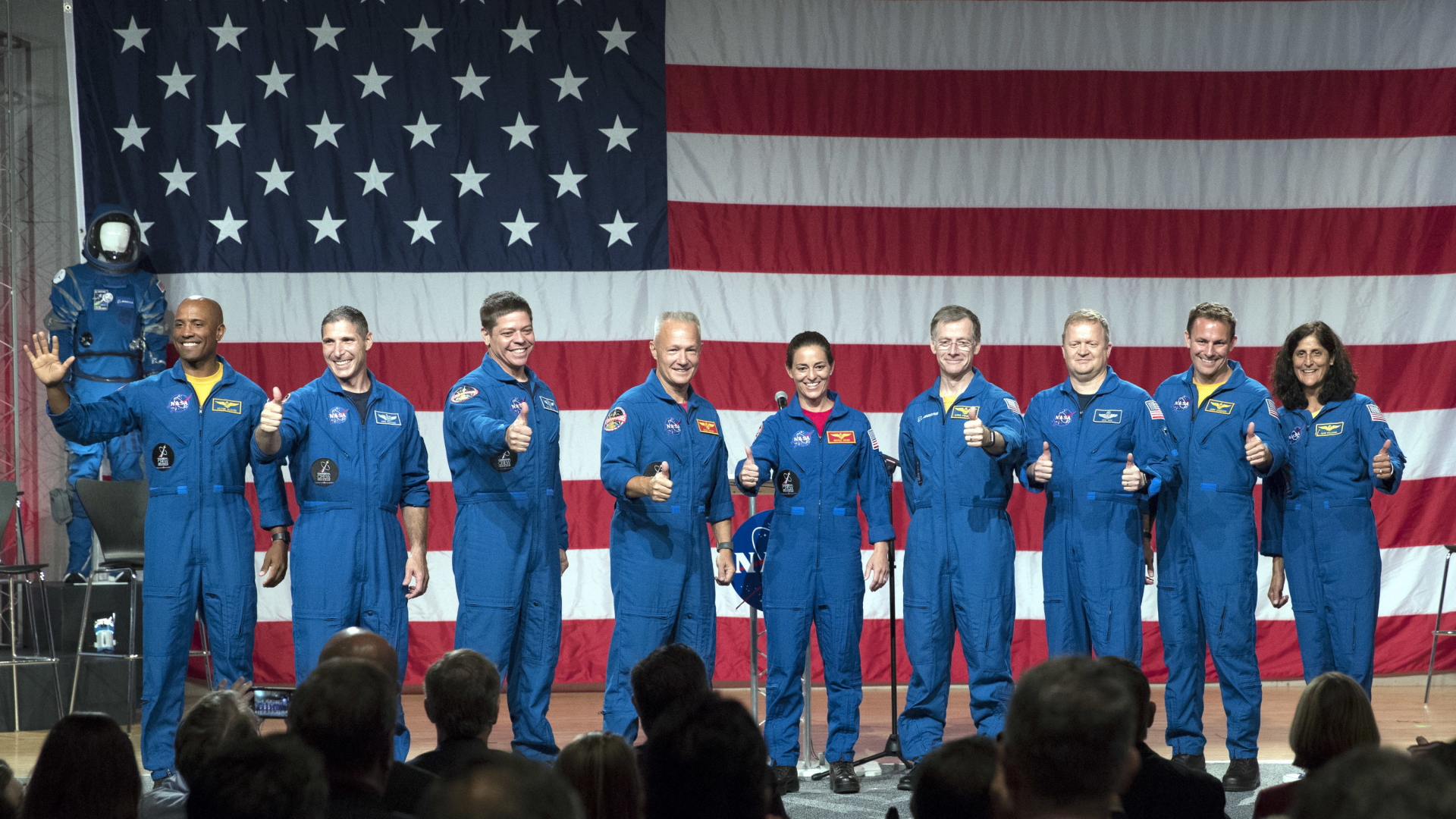 NASA-Astronauten und Astronautinnen heben den Daumen vor einer amerikanischen Flagge.