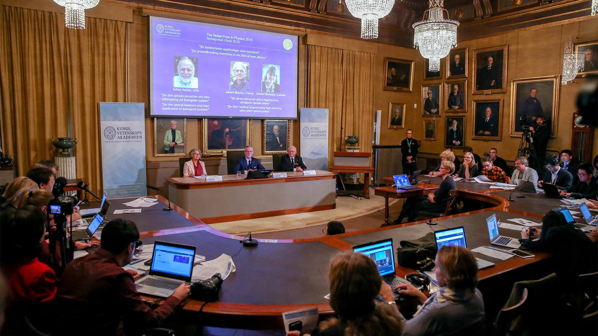 tockholm: Die Nobelpreisträger für Physik 2018, Arthur Ashkin aus den USA (l-r), Gerard Mourou aus Frankreich und Donna Strickland aus Kanada, sind auf einem Bildschirm während der Verkündung in der Königlich Schwedischen Akademie der Wissenschaften zu sehen. | Bildquelle: dpa