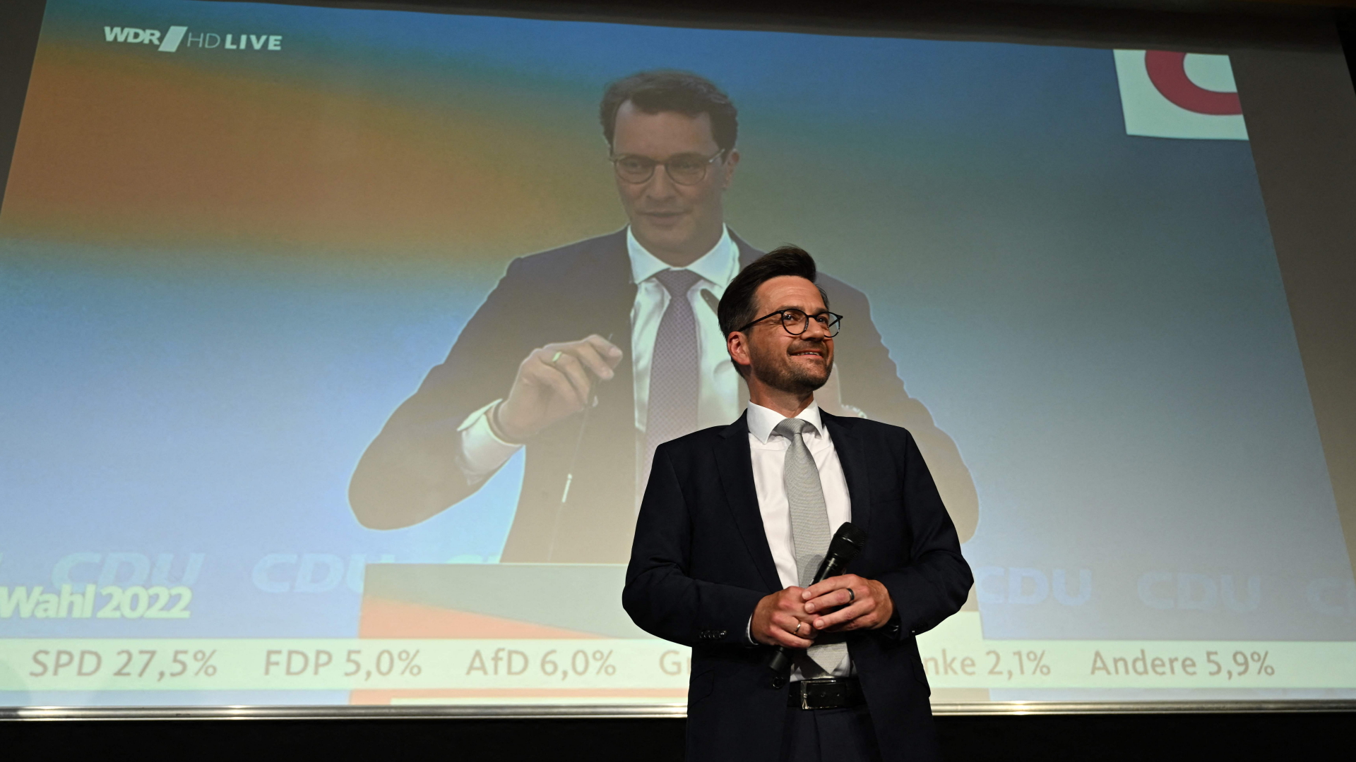 SPD-Herausforderer spricht am Wahlabend zu SPD-Anhängern. Im Hintergrund: Wahlsieger Hendrik Wüst auf einem übergroßen Bildschirm.
