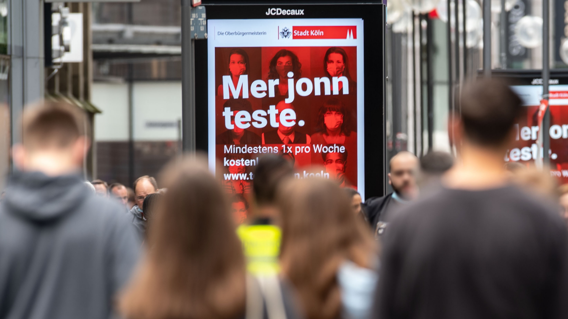 Menschen laufen in Köln auf der Schildergasse an Werbedisplays vorbei, auf denen "Mer jonn teste" (Kölsch für "Wir gehen testen") steht.