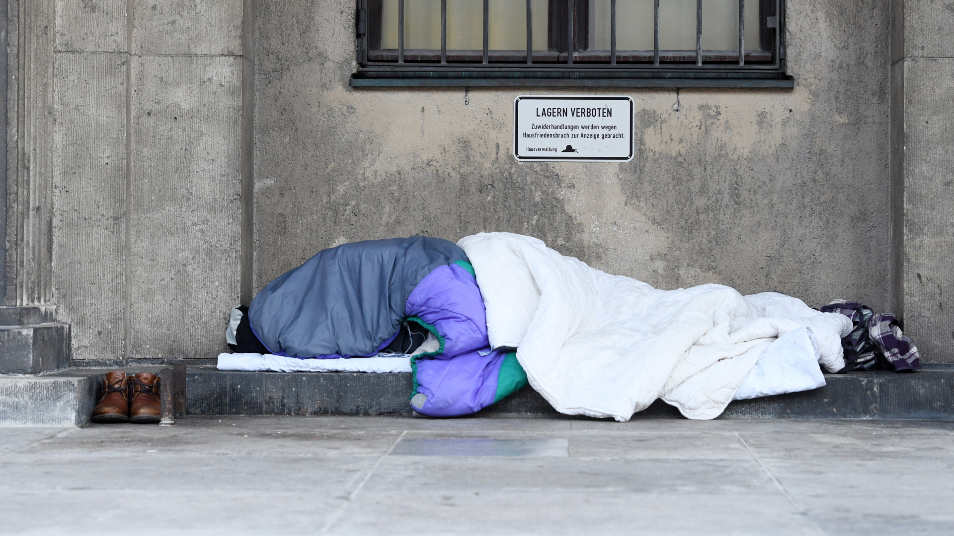 Ein Obdachloser liegt, eingehüllt in Decken und einem Schlafsack, vor einem Gebäude in München. | Bildquelle: dpa