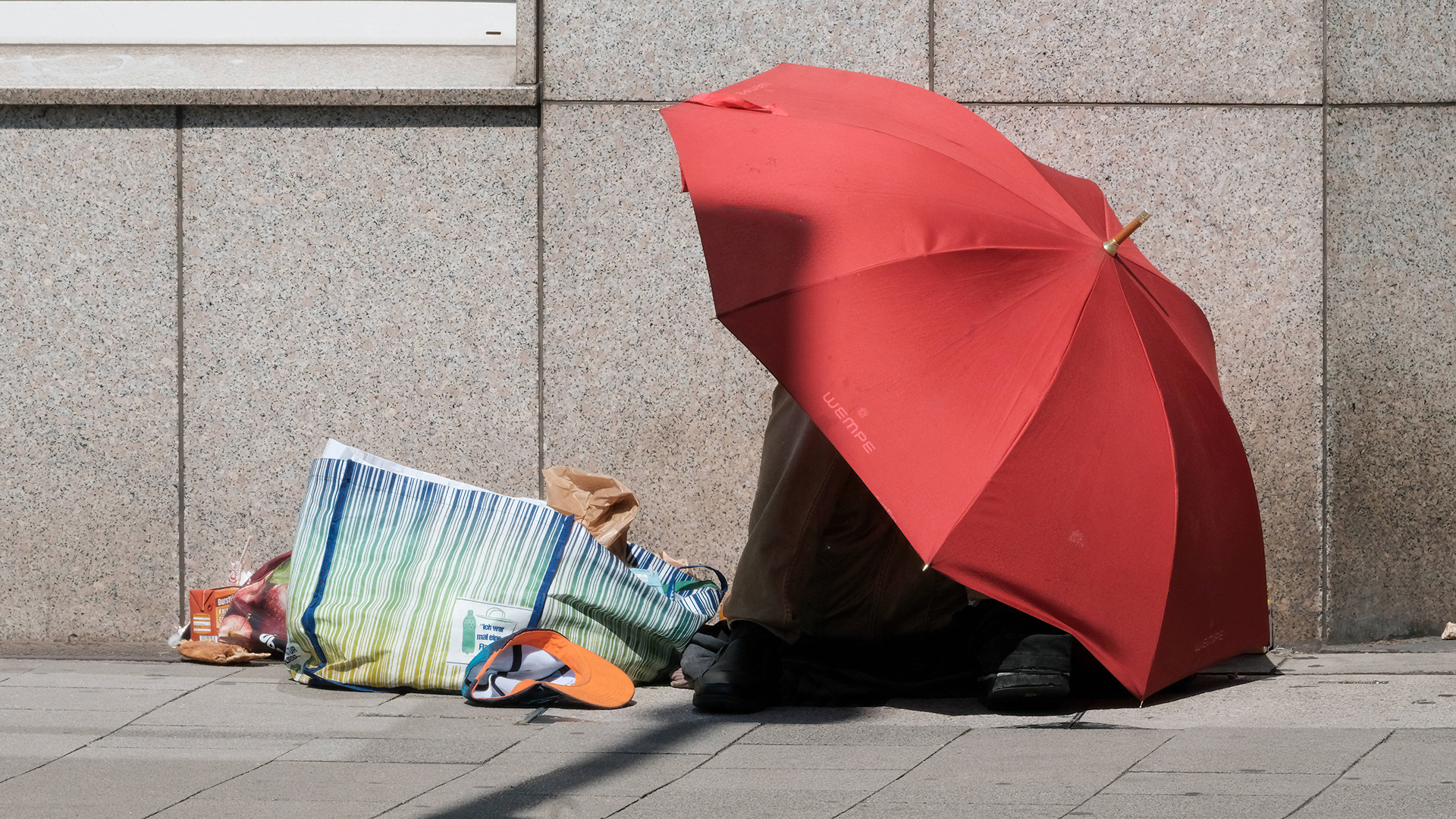 Ein Obdachloser sitzt unter einem Schirm auf der Straße. | IMAGO/Michael Gstettenbauer