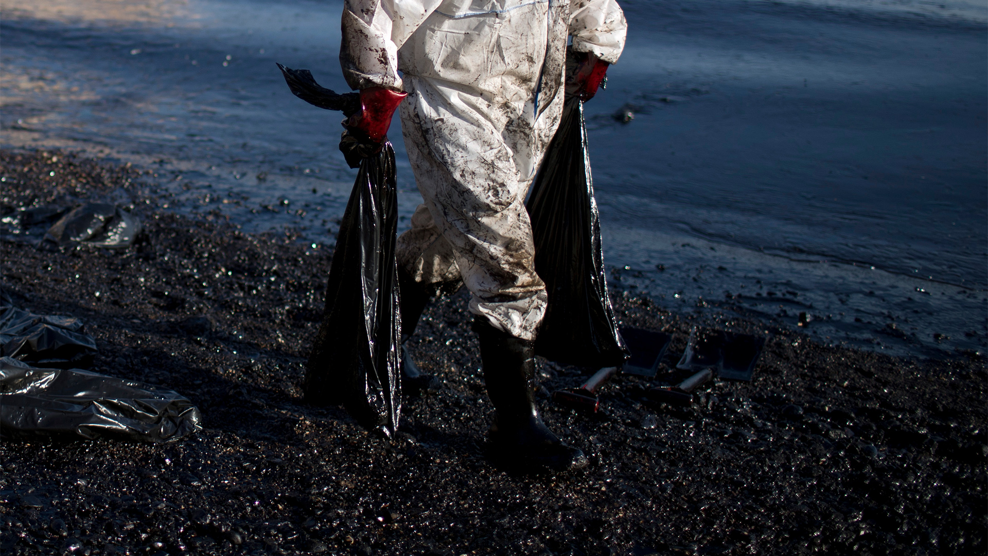 Ein Mann in Schutzanzug trägt Plastikbeutel an einem mit Öl verschmutzten Strand.
